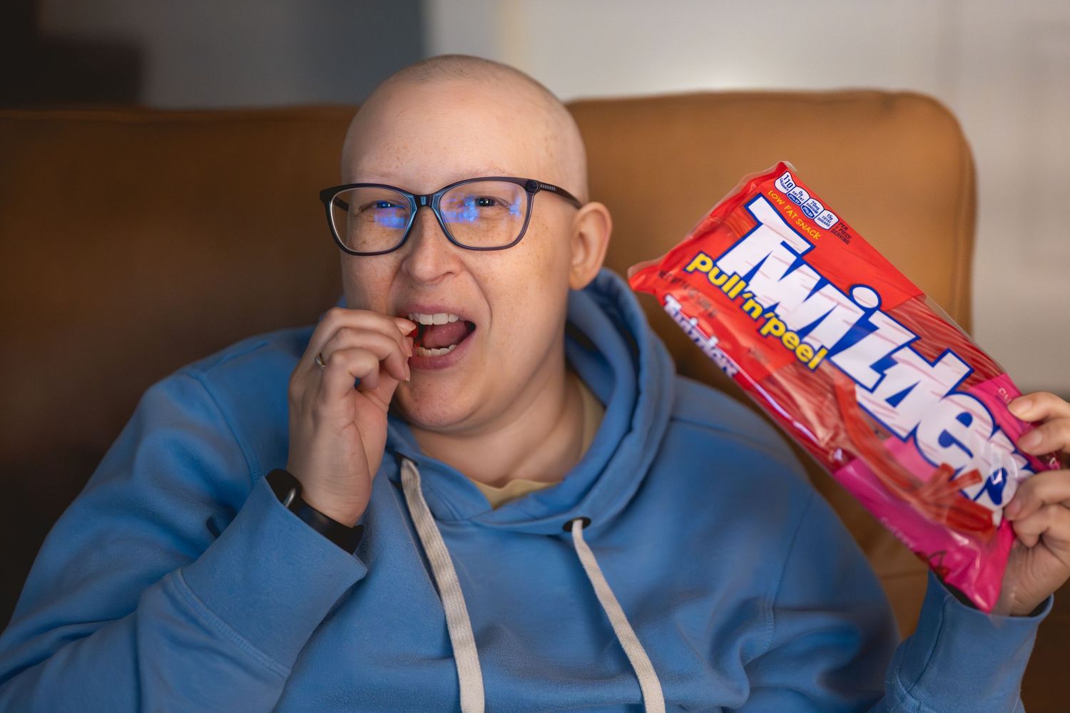 Bald woman with glasses smiling while eating Twizzlers candy, wearing a blue hoodie, seated in a cozy chair.