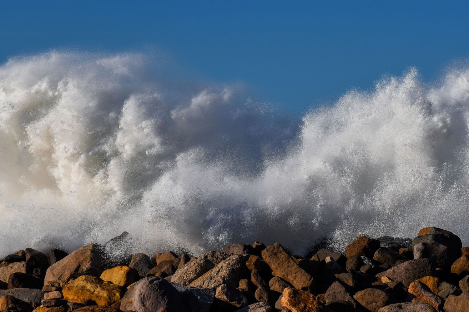 Massive ocean waves crashing over rocky shoreline with white sea spray against a bright blue sky.