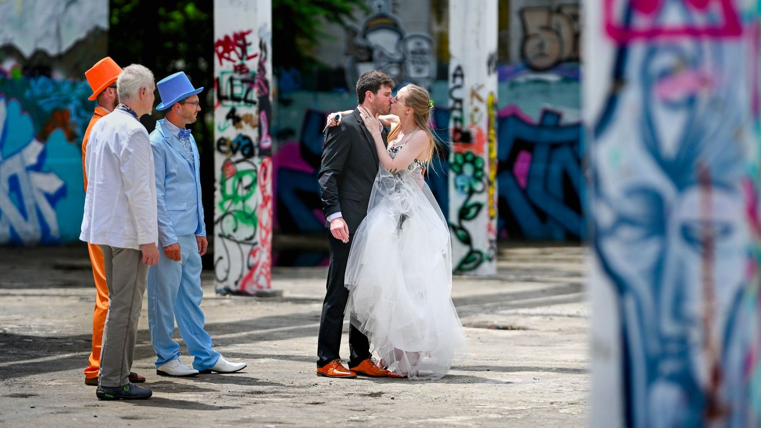A couple poses in front of a colorful graffiti wall on their wedding day while wearing formal attire.