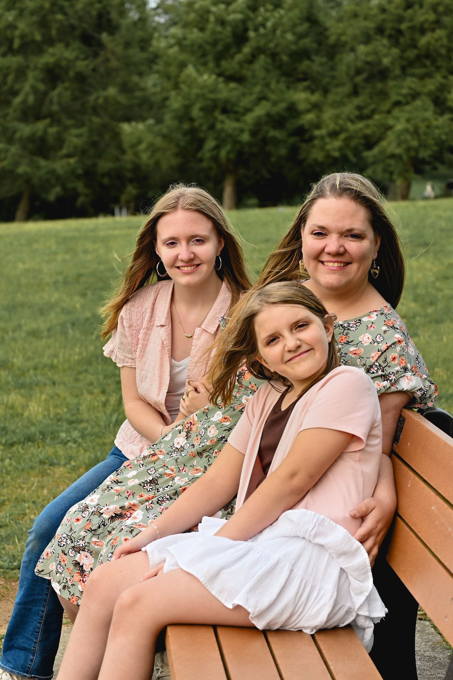 Three smiling females pose together on a wooden park bench surrounded by lush green trees and grass.