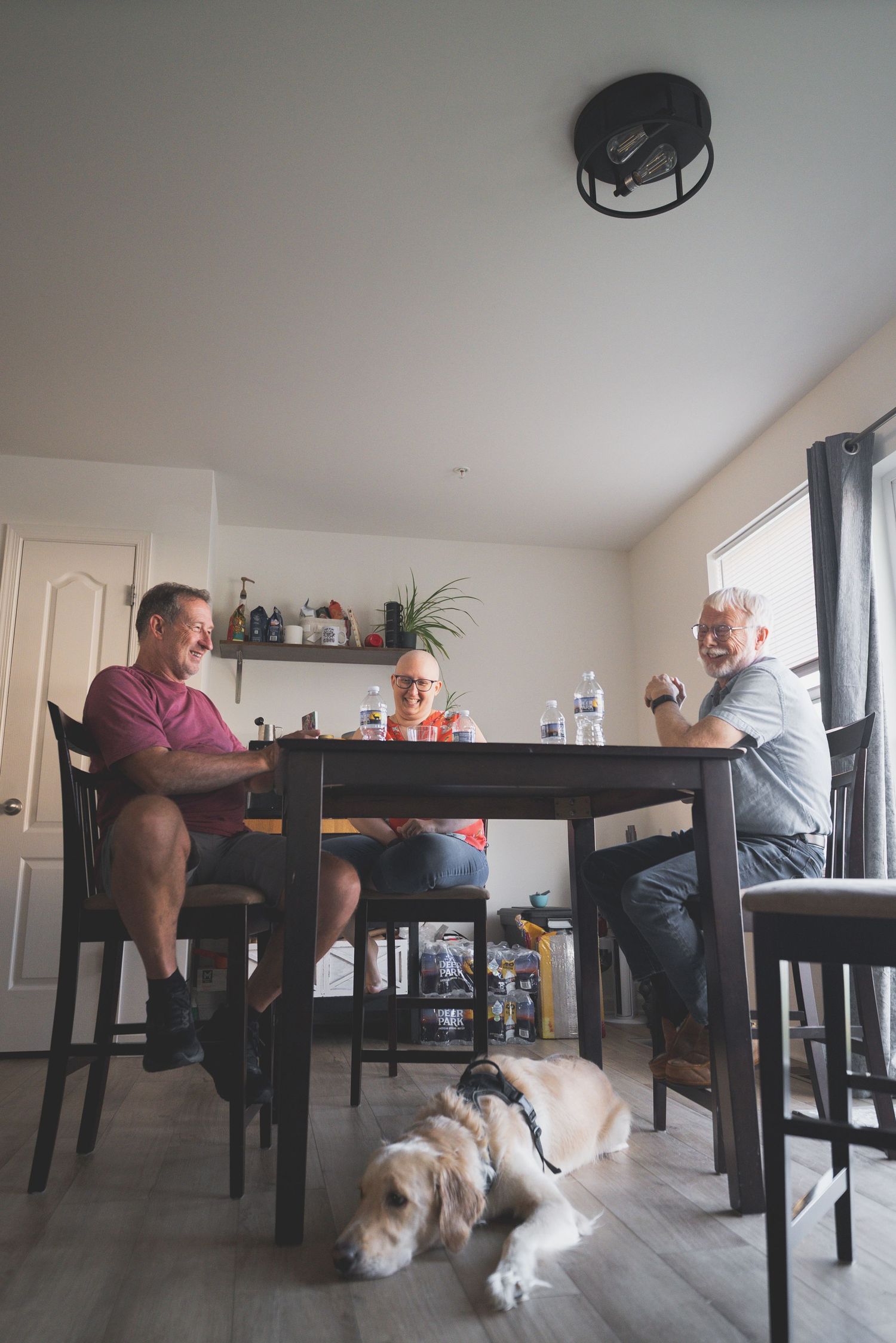 Three men sit around a dark wooden dining table chatting, with a golden dog resting on the floor beneath them.
