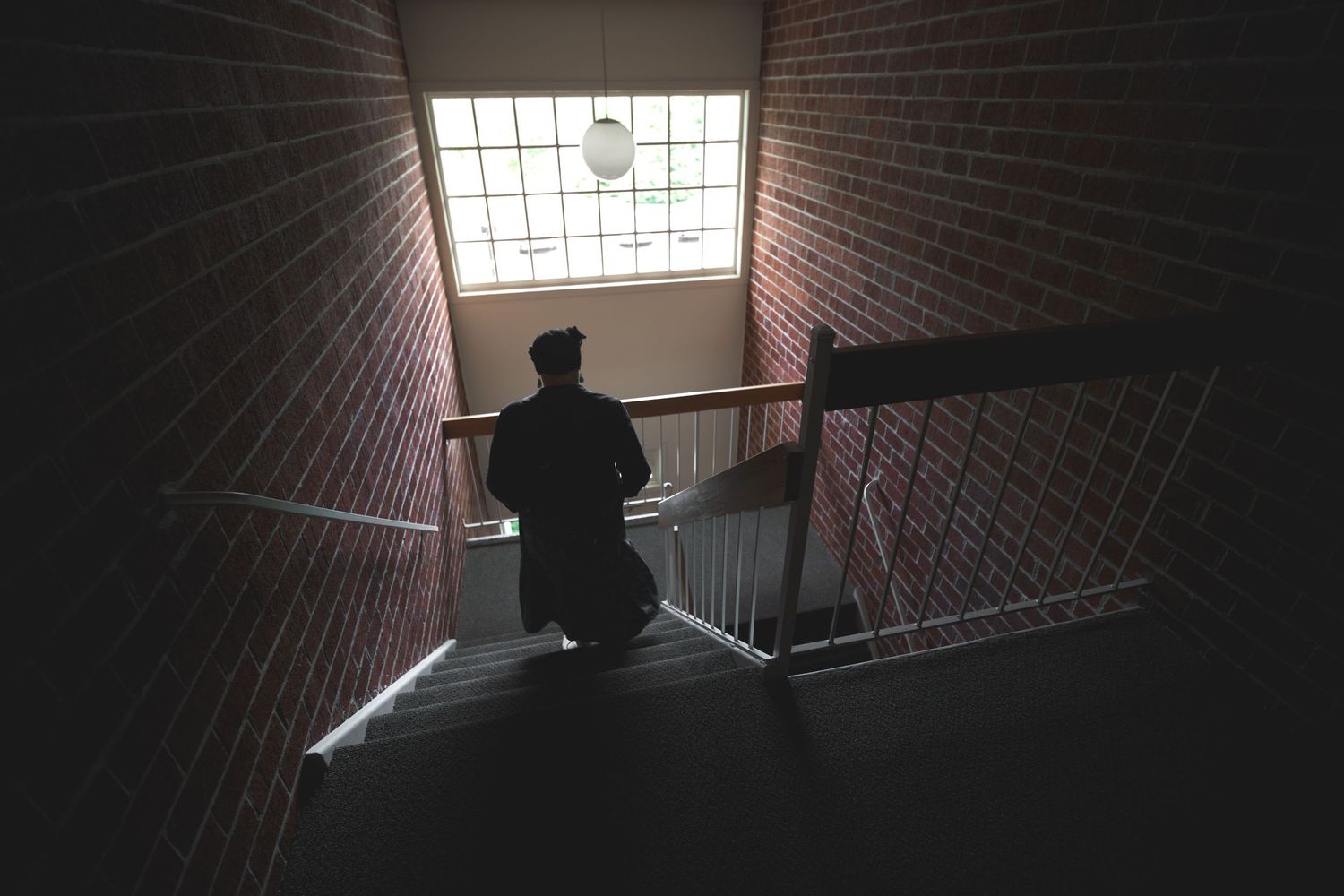 Silhouetted person standing on dark staircase with brick walls and natural light from window above.