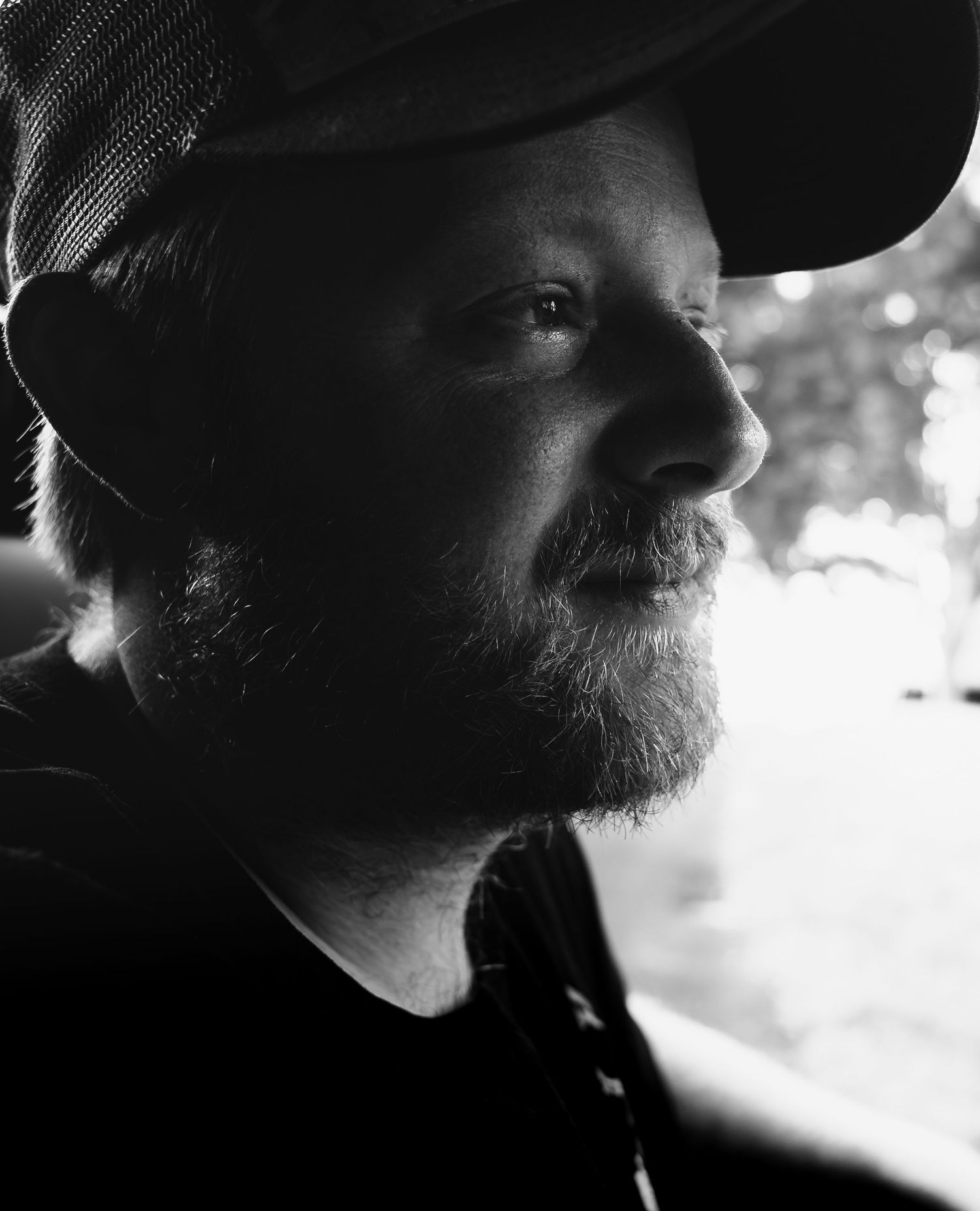 Black and white portrait of a bearded man wearing a cap, captured in a moody side profile with soft bokeh background.