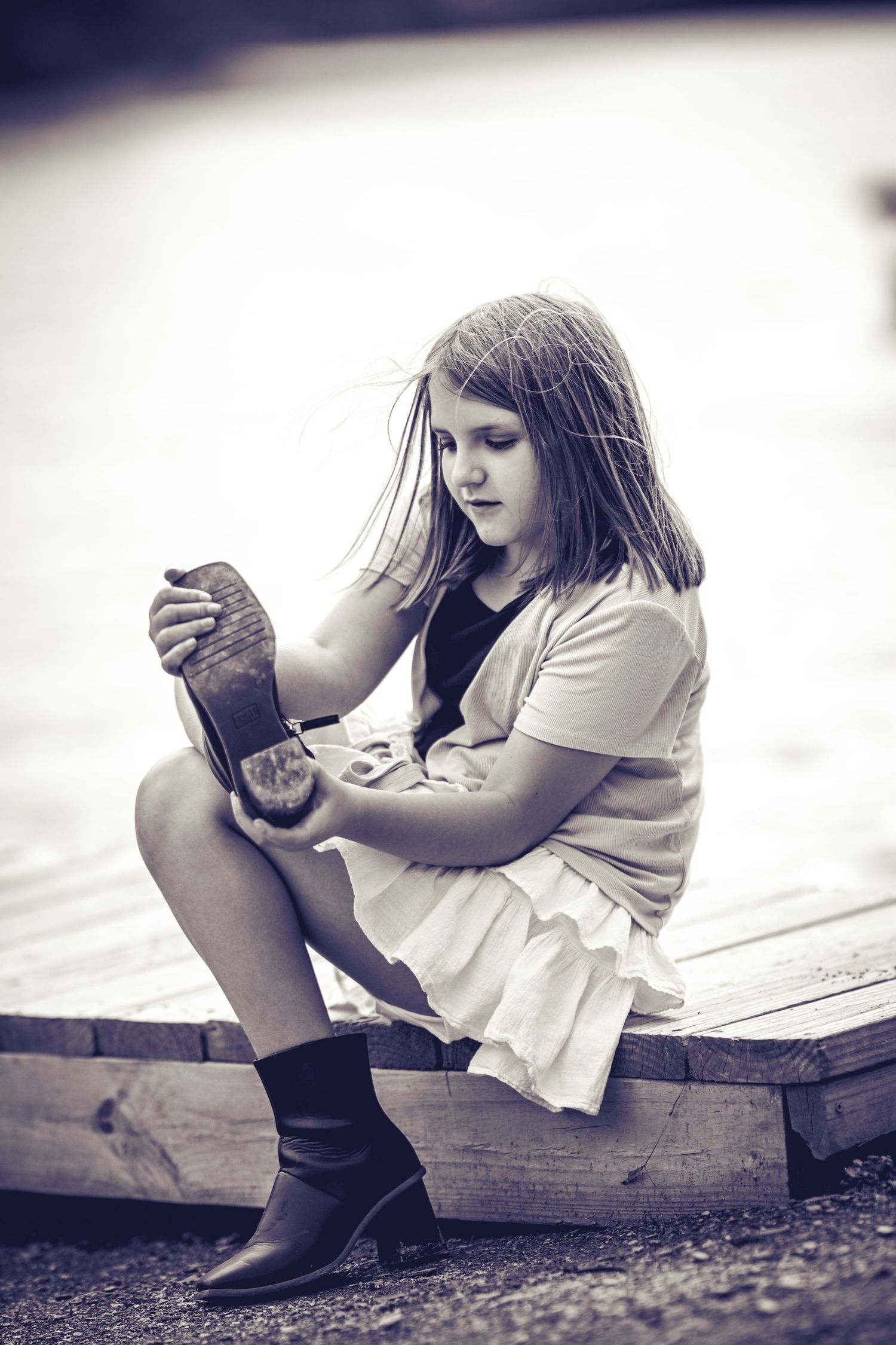 Young girl in boots and light dress sits on wooden surface while examining something in black and white photo.