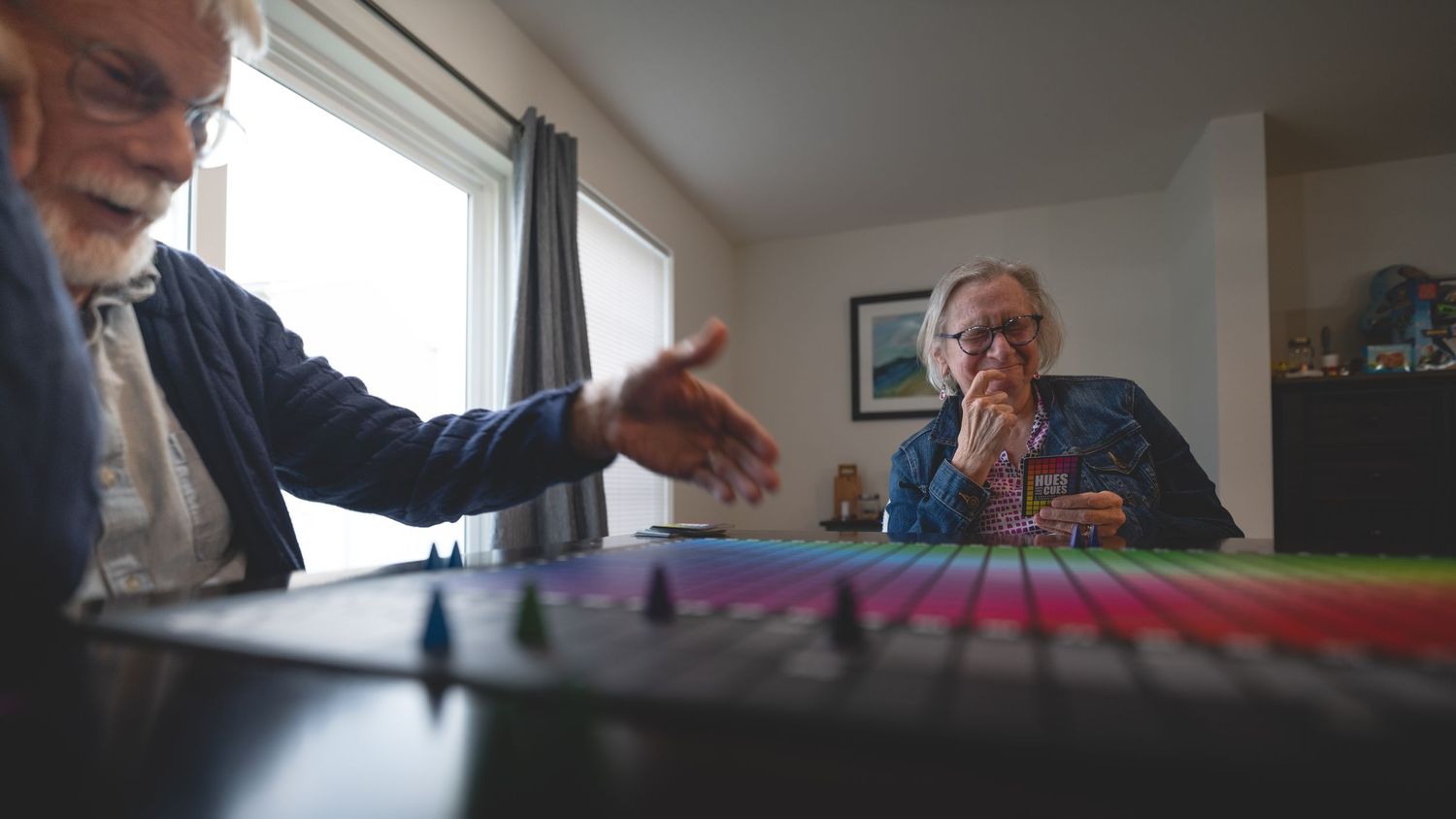 Two elderly people playing a colorful board game indoors, one reaching across the table in a bright living room.