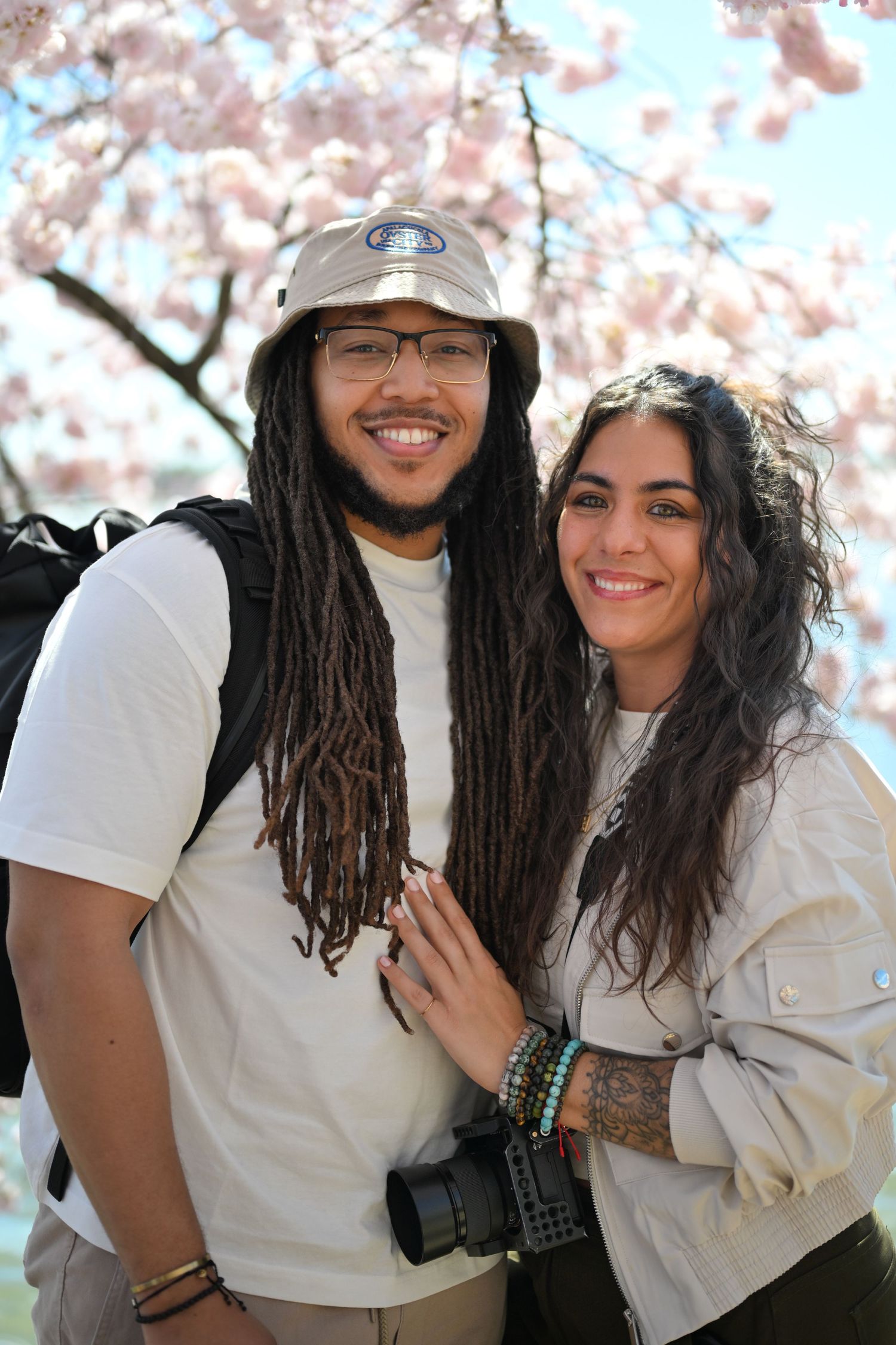 A couple poses under blossoming cherry trees with one holding a camera, both wearing white tops on a sunny spring day.