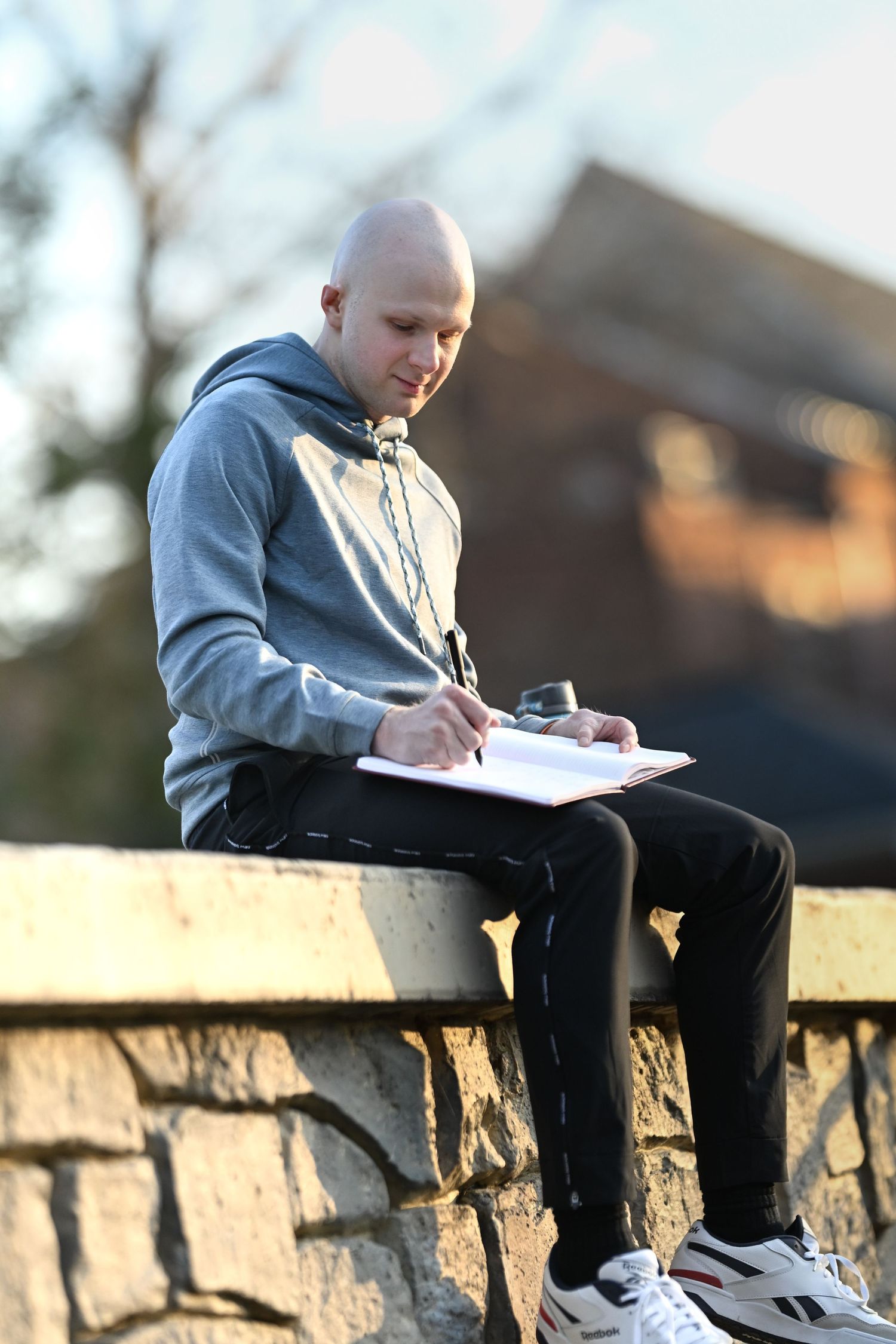 Person in gray hoodie writing in notebook while sitting on stone wall.
