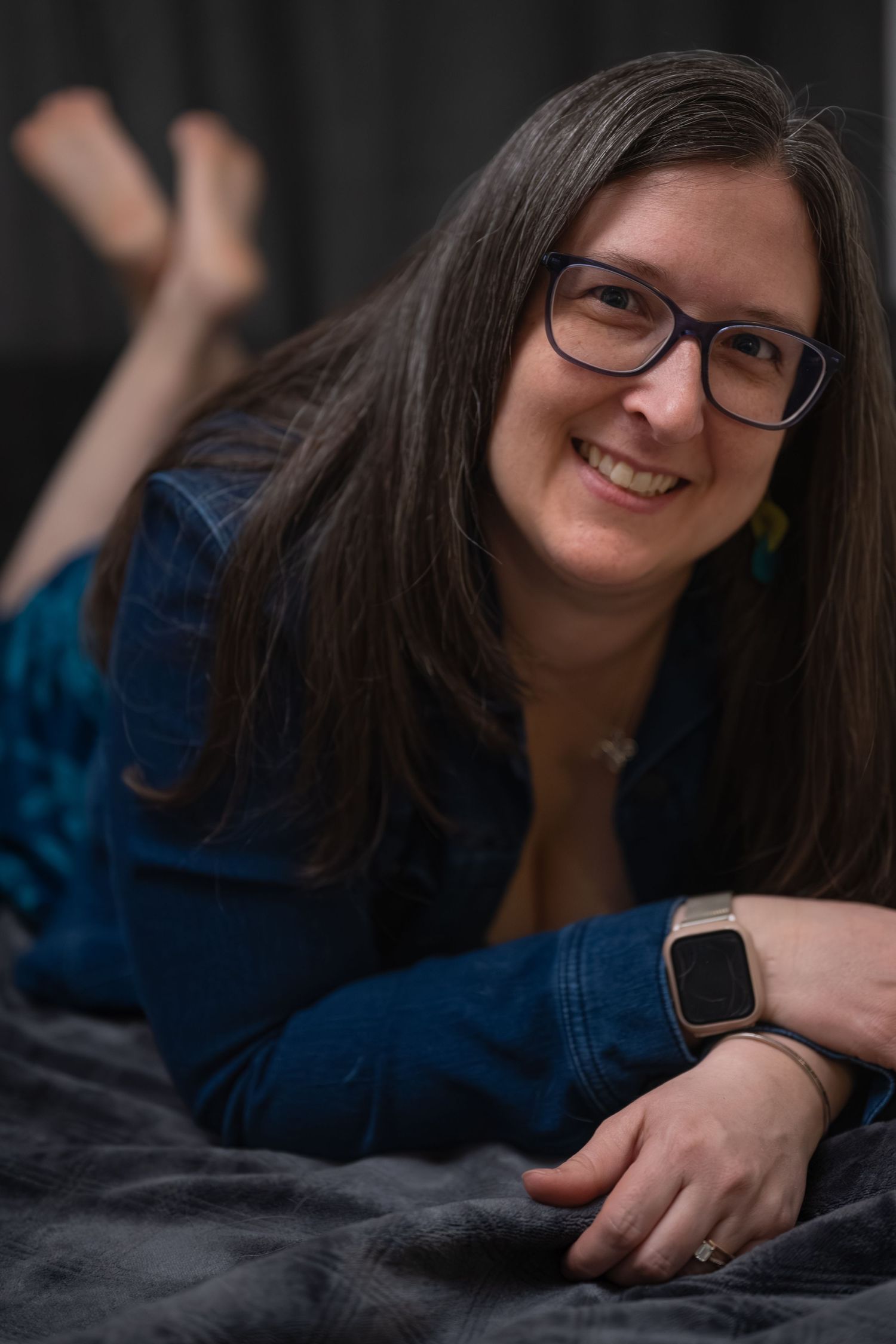 Person with glasses and long dark hair wearing a blue top poses playfully while lying down against a dark background.