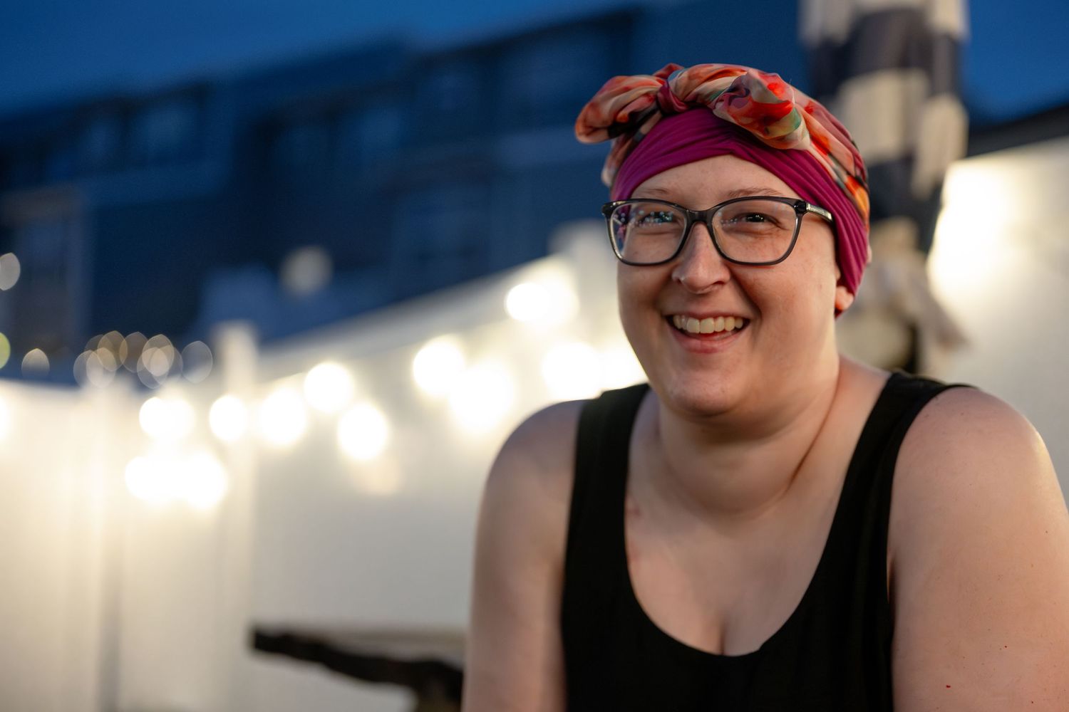 Smiling woman wearing colorful head wrap and glasses outdoors at dusk with string lights in background.