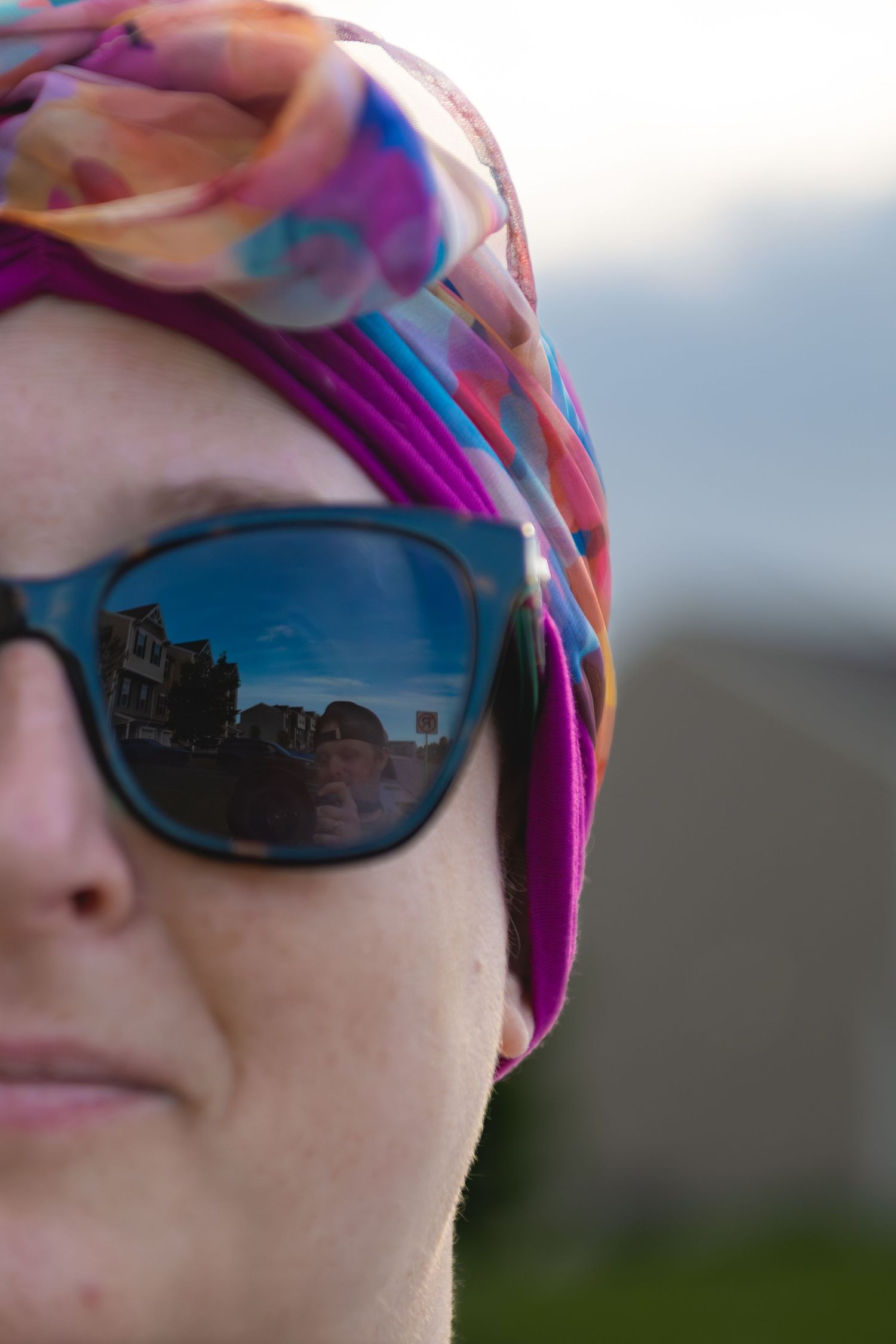 Woman wearing colorful silk headscarf and sunglasses reflecting a street scene outdoors.