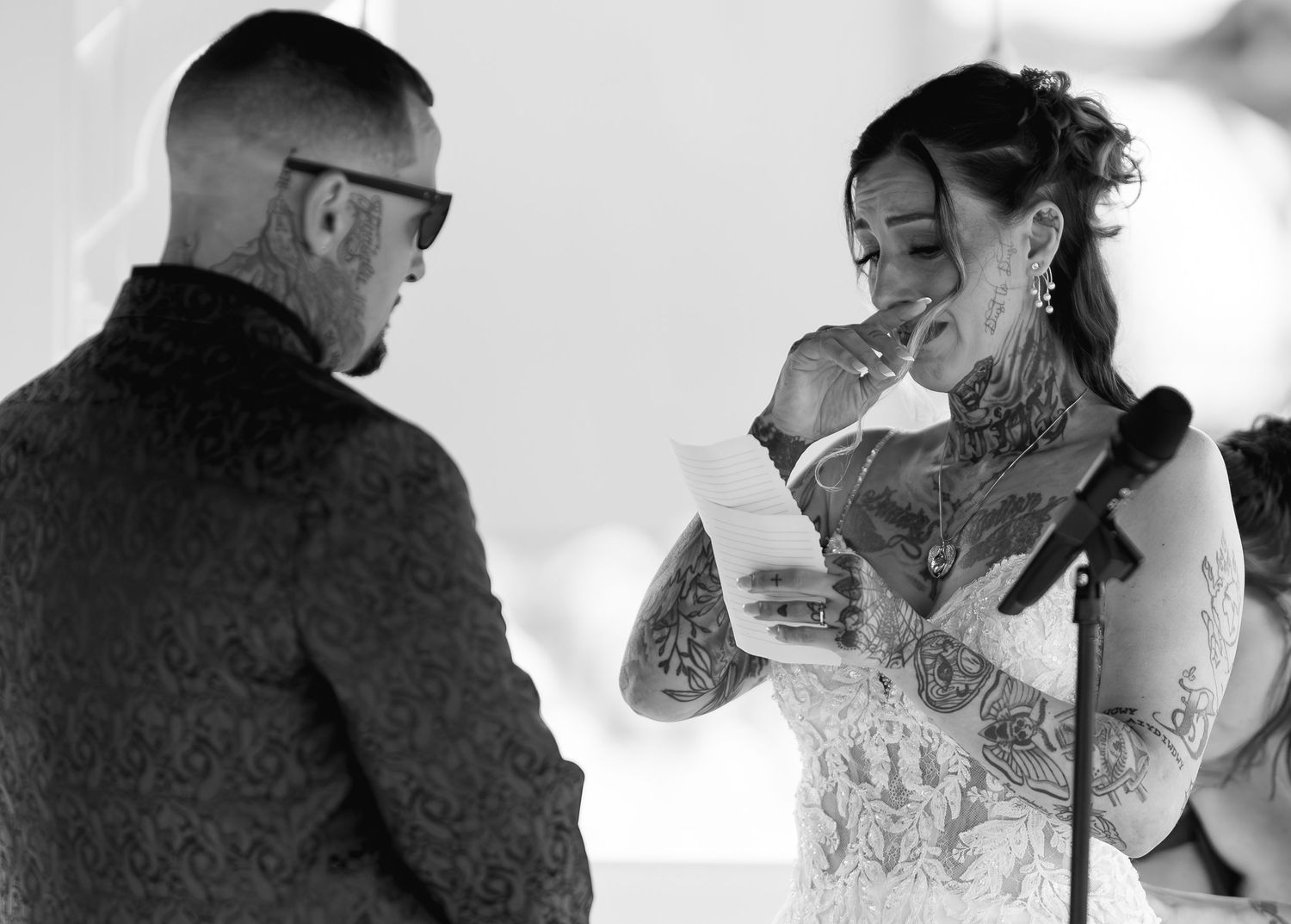 Black and white wedding photo of tattooed bride reading vows while emotional groom listens during outdoor ceremony.