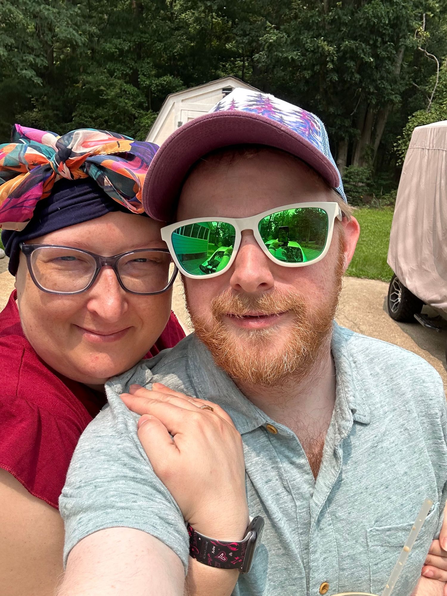 A smiling couple takes a selfie outdoors, woman wearing colorful headscarf and glasses, man in maroon cap and green sunglasses.