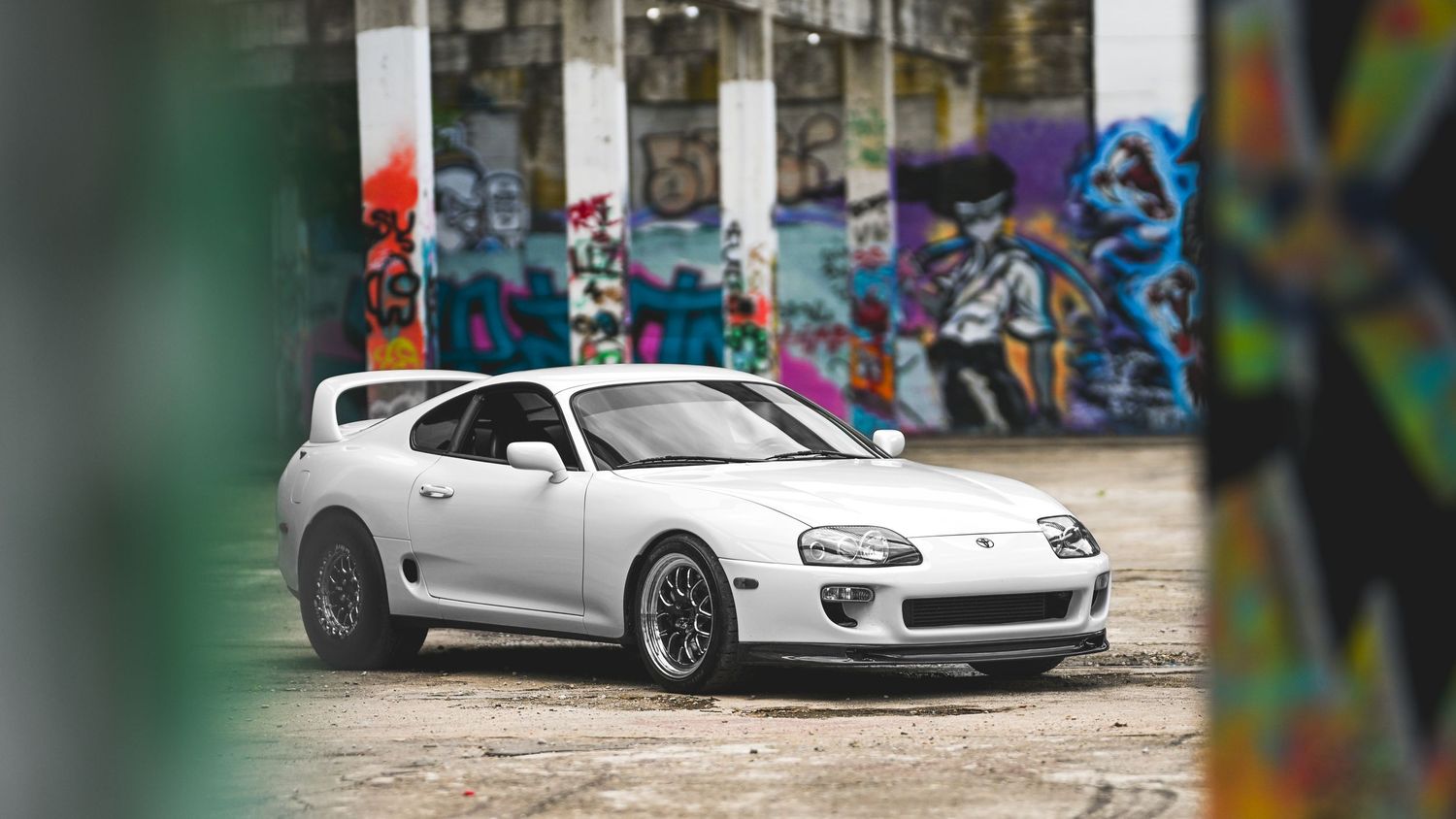 A silver Toyota Supra MK4 sports car parked in front of a graffiti-covered wall with colorful street art.