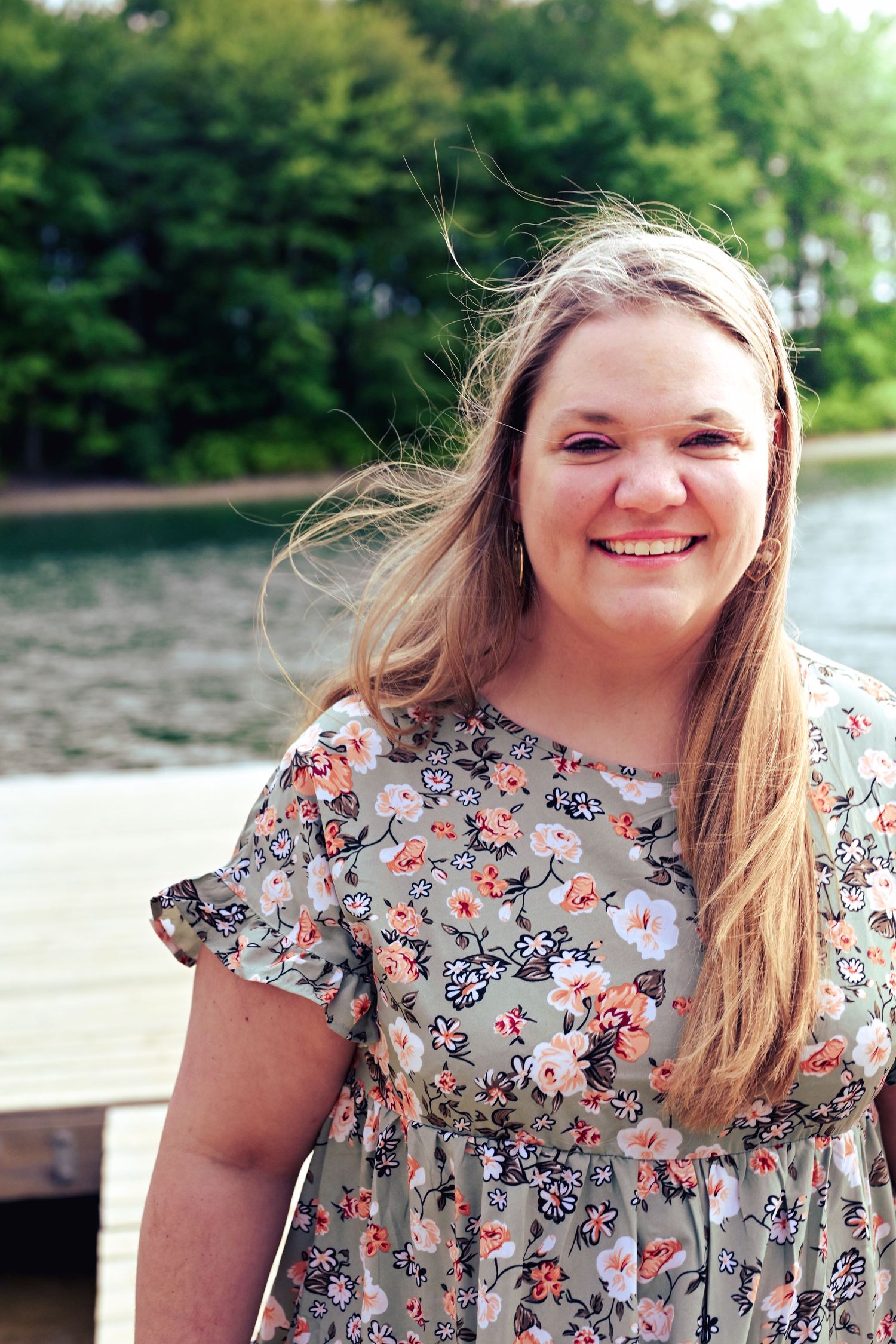A person in a floral print dress stands by a waterfront on a sunny day, with trees visible in the background.