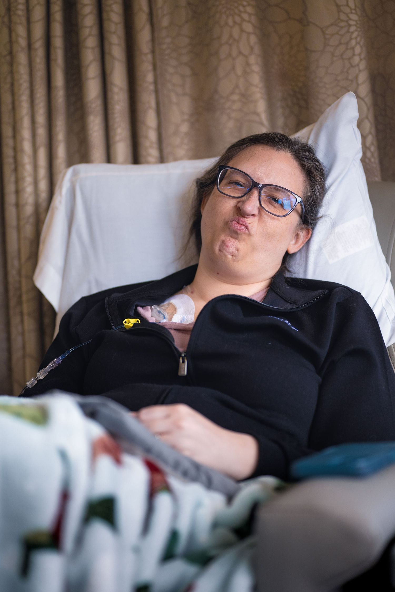 Woman with glasses resting in hospital bed, making a funny face while covered with a colorful blanket.