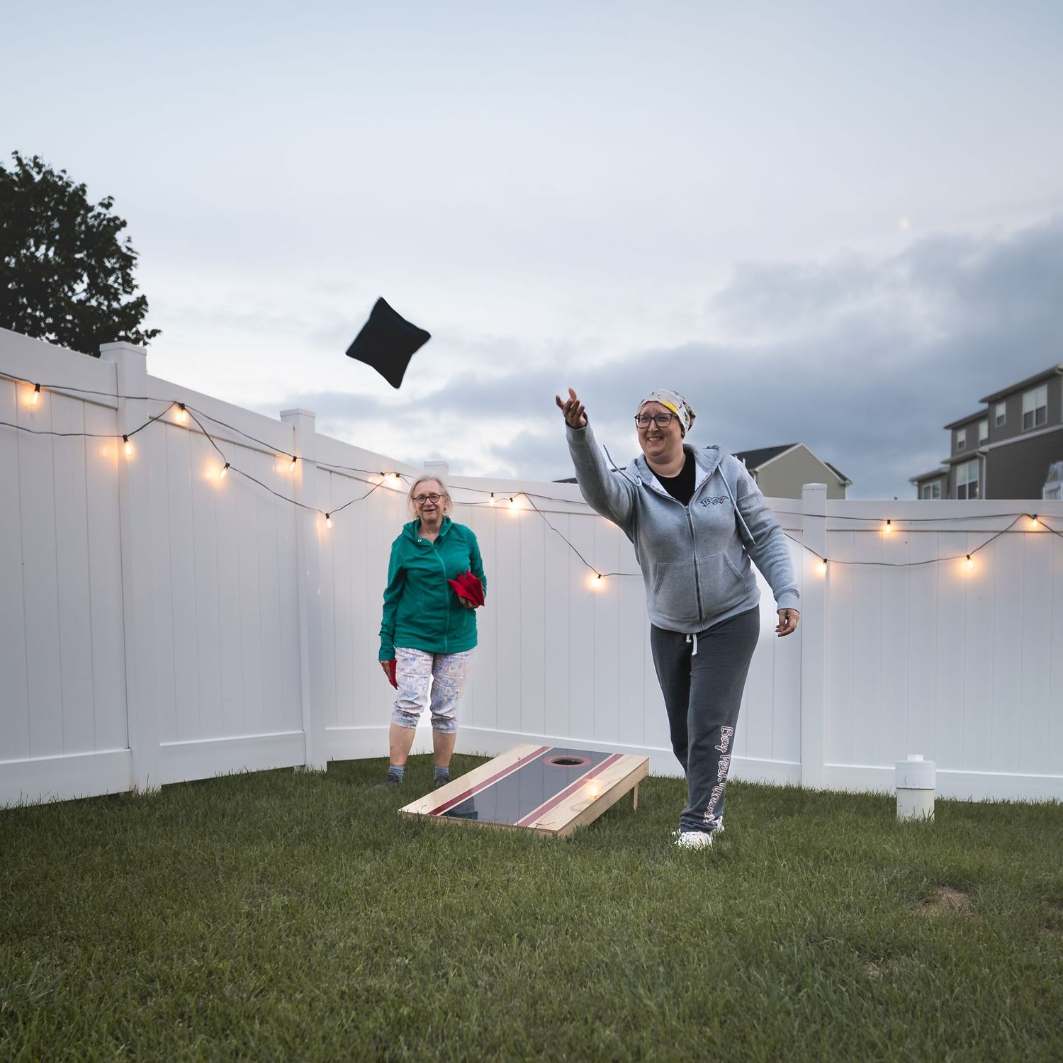 Two women playing cornhole in a backyard with string lights on a white fence at dusk.