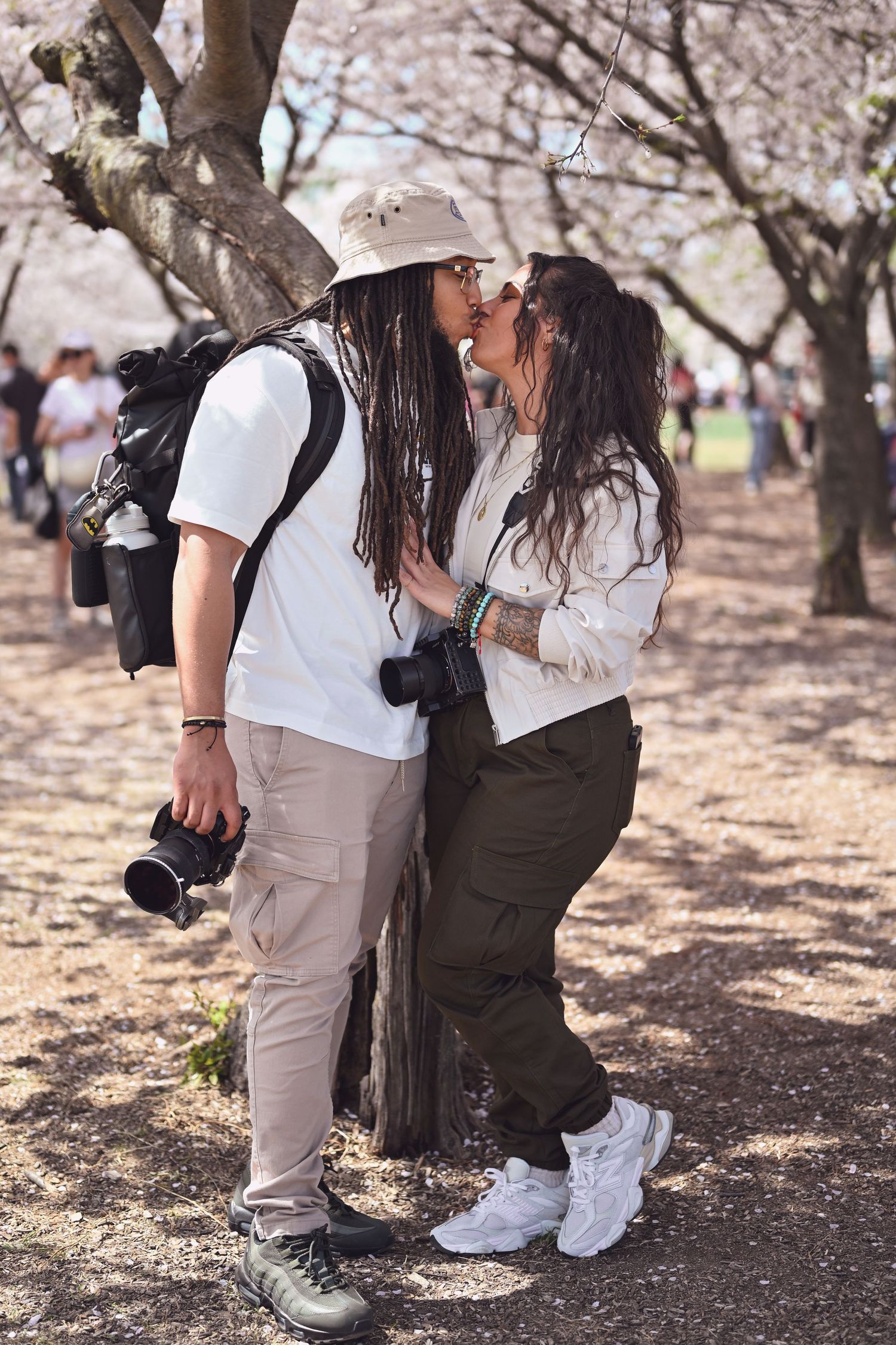 A couple shares an intimate moment amongst blooming cherry blossom trees while carrying photography equipment.