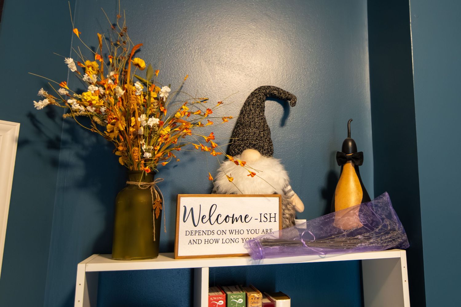 Cozy shelf decor with gnome figurine, Welcome-ish sign, fall floral arrangement in gold vase against teal wall.