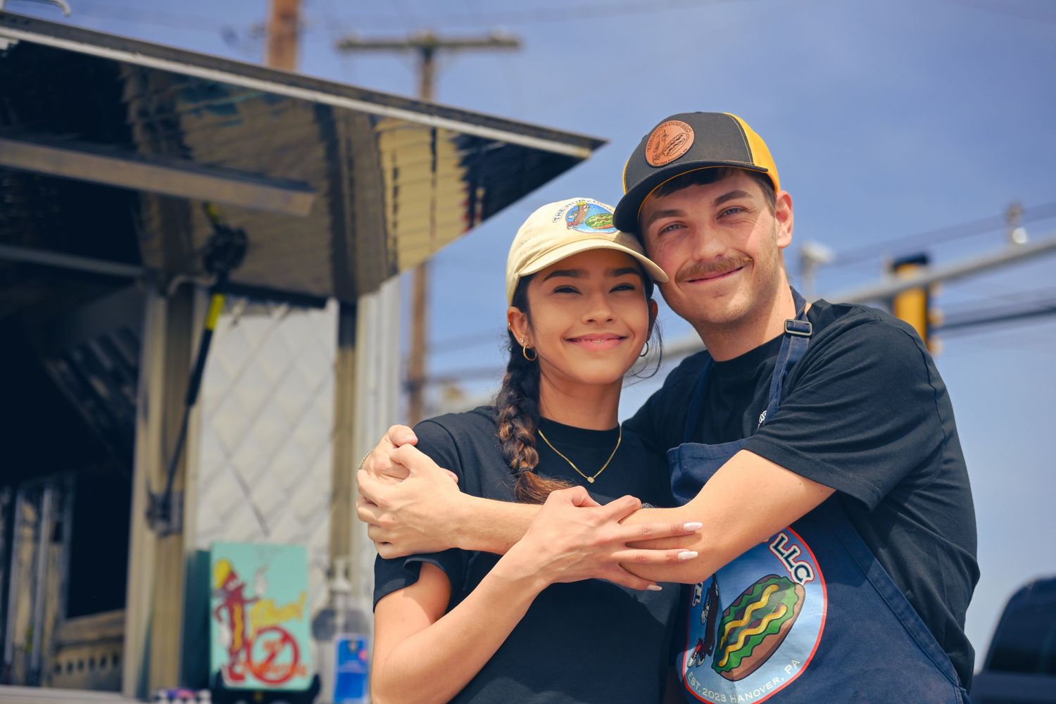 Two people hugging and smiling while wearing baseball caps and casual clothing outdoors.