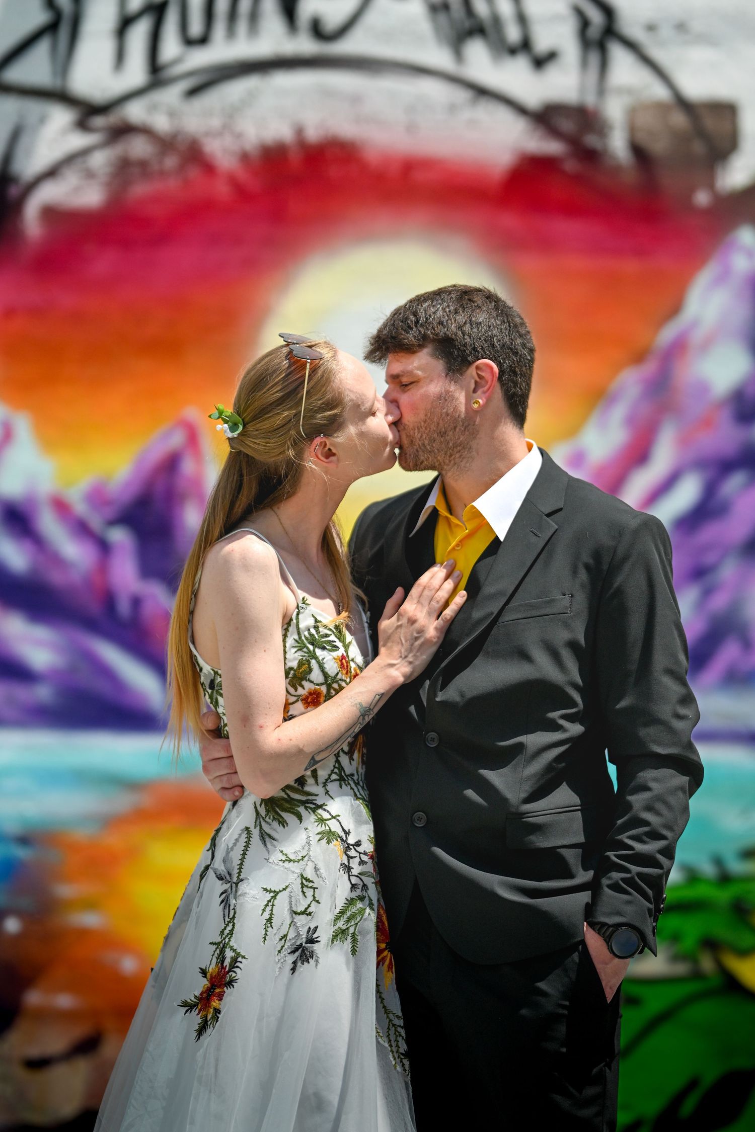 A couple shares a romantic kiss against a vibrant street art mural featuring mountains and a sunset in rainbow colors.