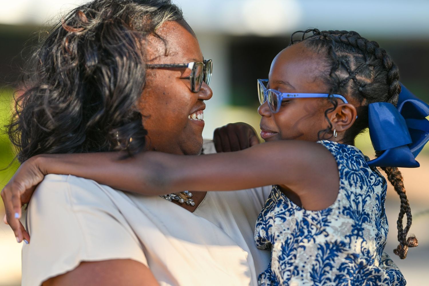 A woman and young girl with matching glasses share a warm embrace and smile outdoors.