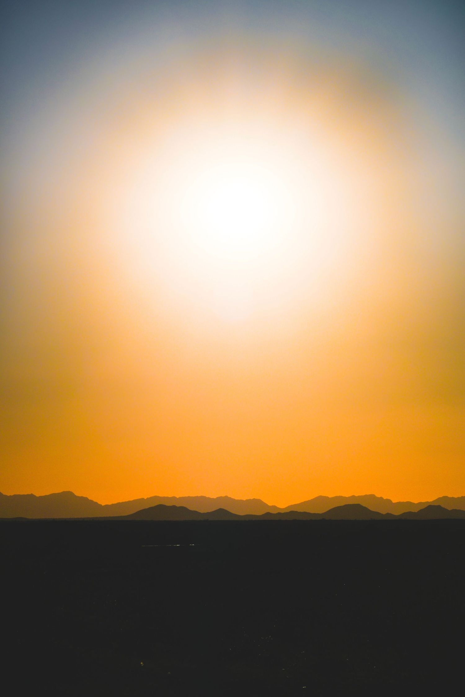Stunning desert sunset with glowing sun, orange sky gradient, and silhouetted mountain range on the horizon.