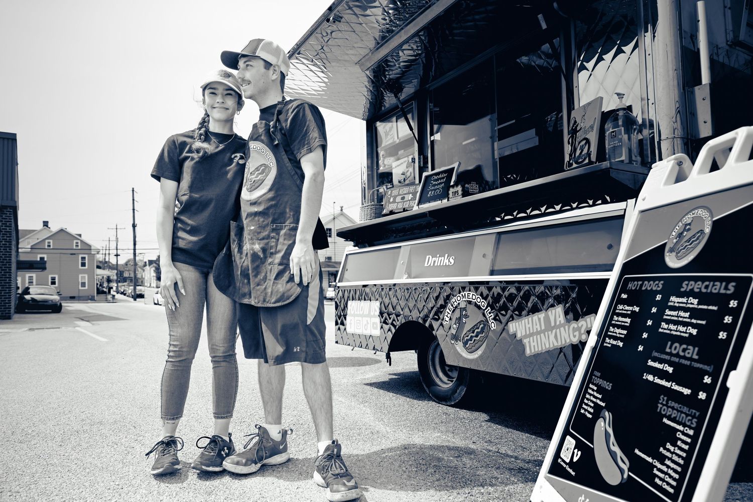 Two people stand outside a food truck with a menu board on the street in a black and white photograph.