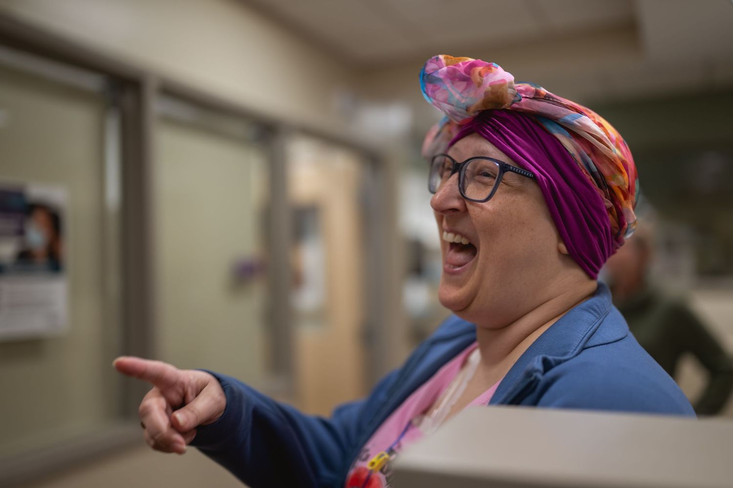 A laughing woman wearing colorful head wrap and glasses points finger in a hospital hallway.