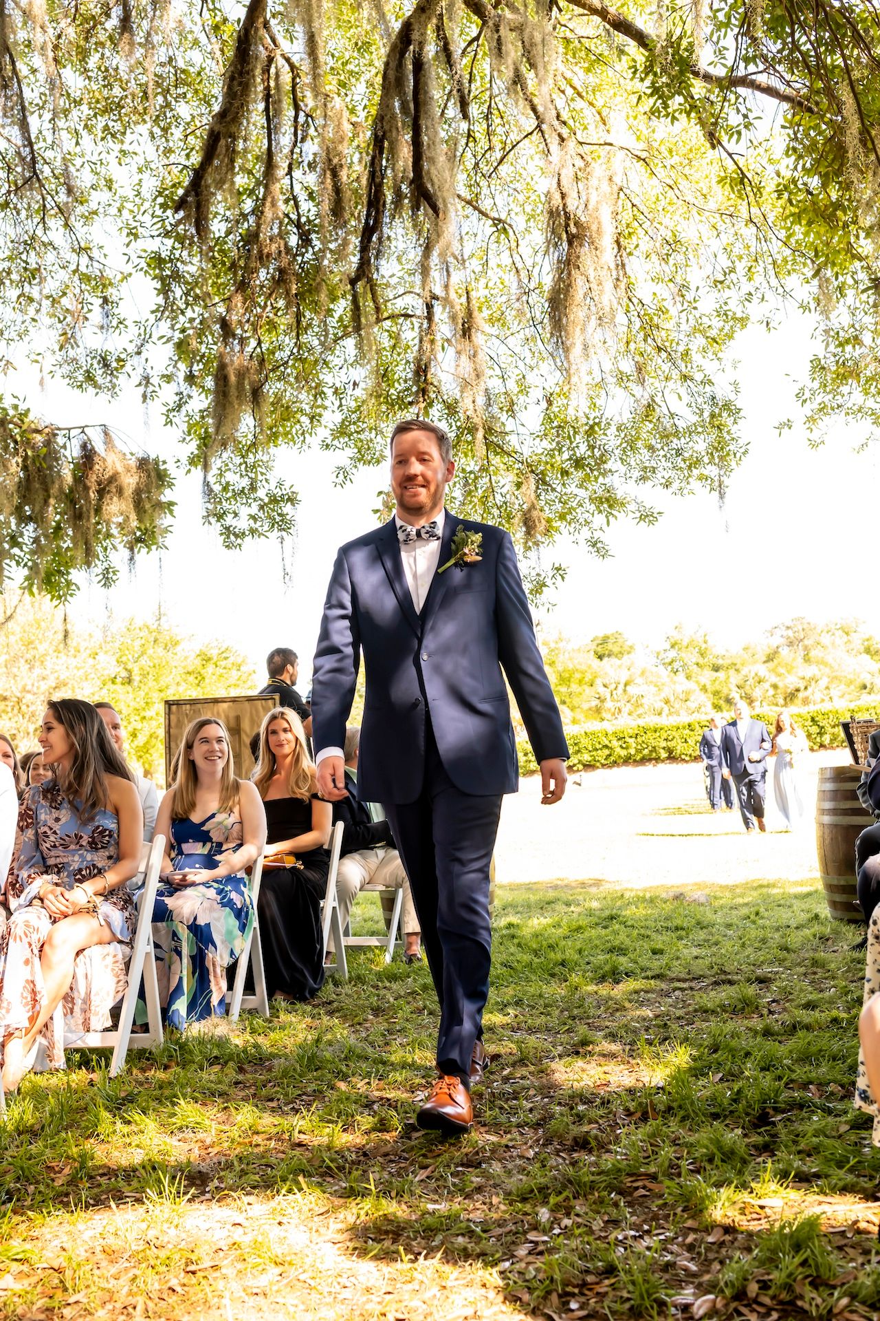 A person in a navy suit walks down an outdoor aisle beneath Spanish moss-draped trees at a wedding ceremony.