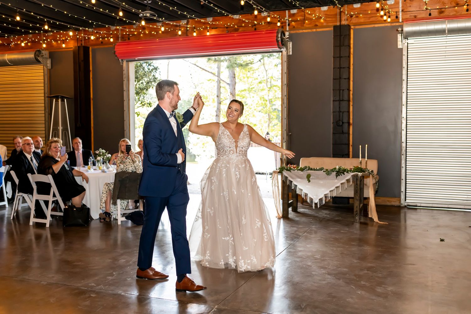 A couple shares their first dance in a rustic venue decorated with twinkling string lights and white table settings.