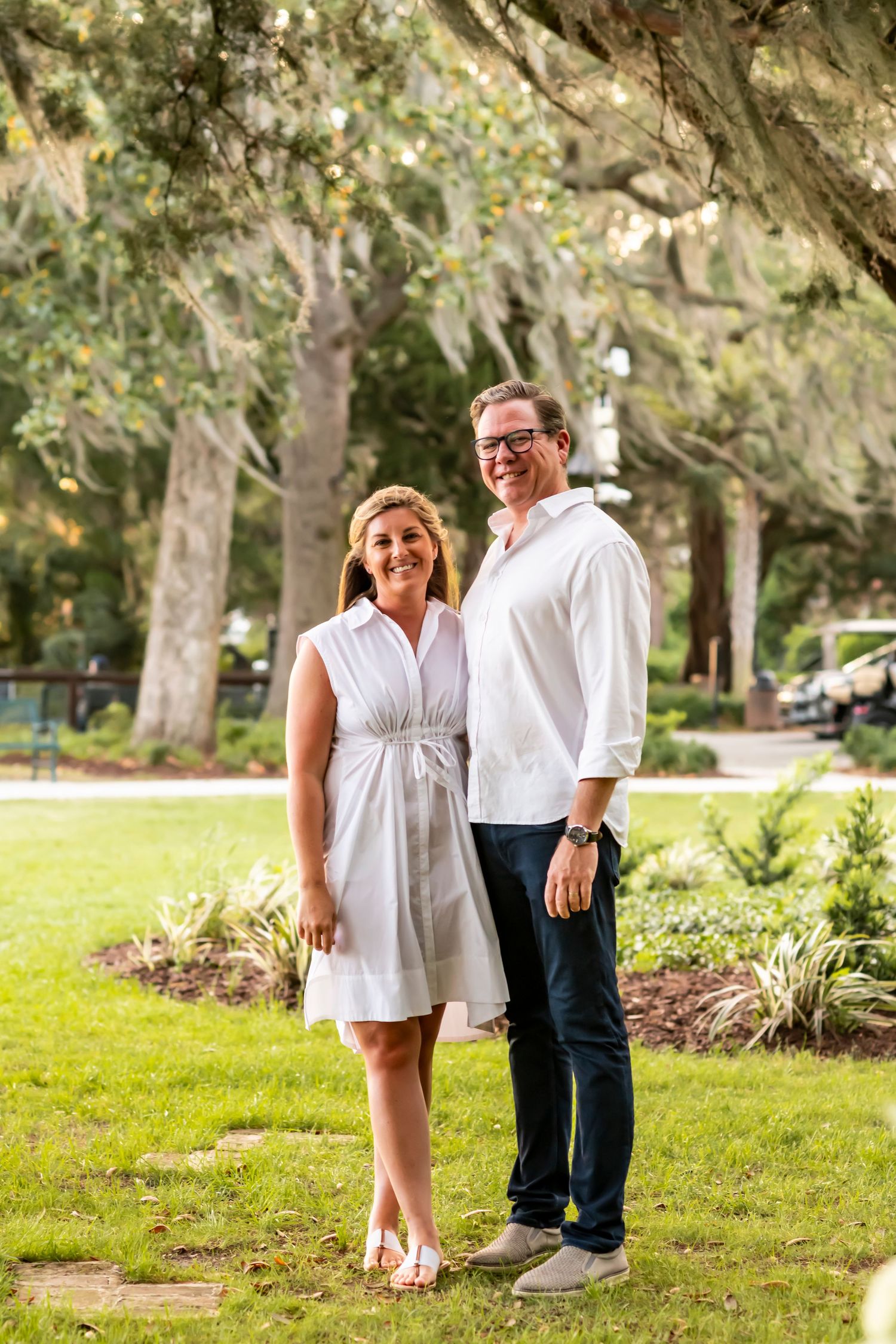 Couple standing together on green lawn in a park with trees and shrubbery in the background on sunny day.