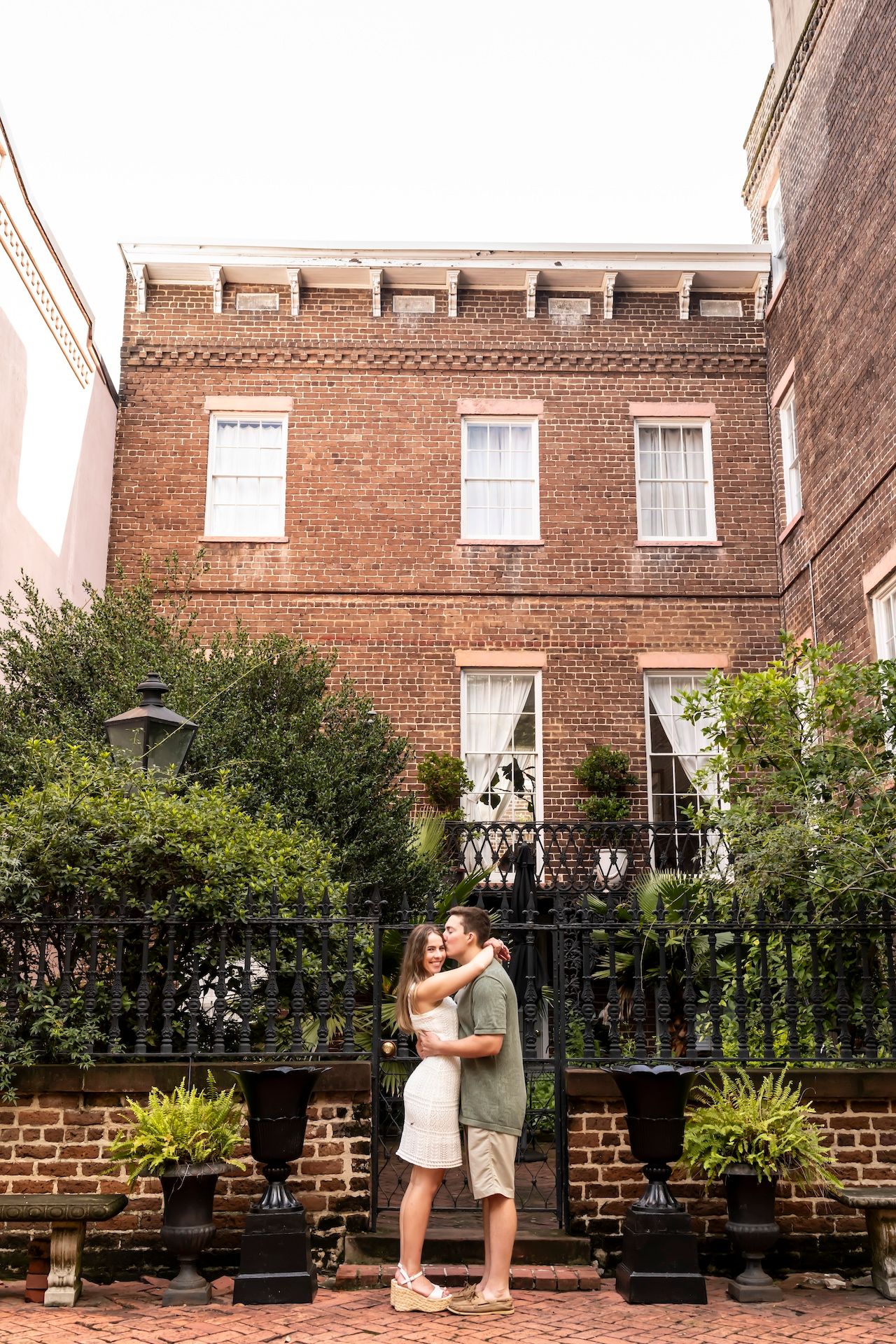 Two people embrace in front of a historic brick building with white windows and lush greenery in planters.