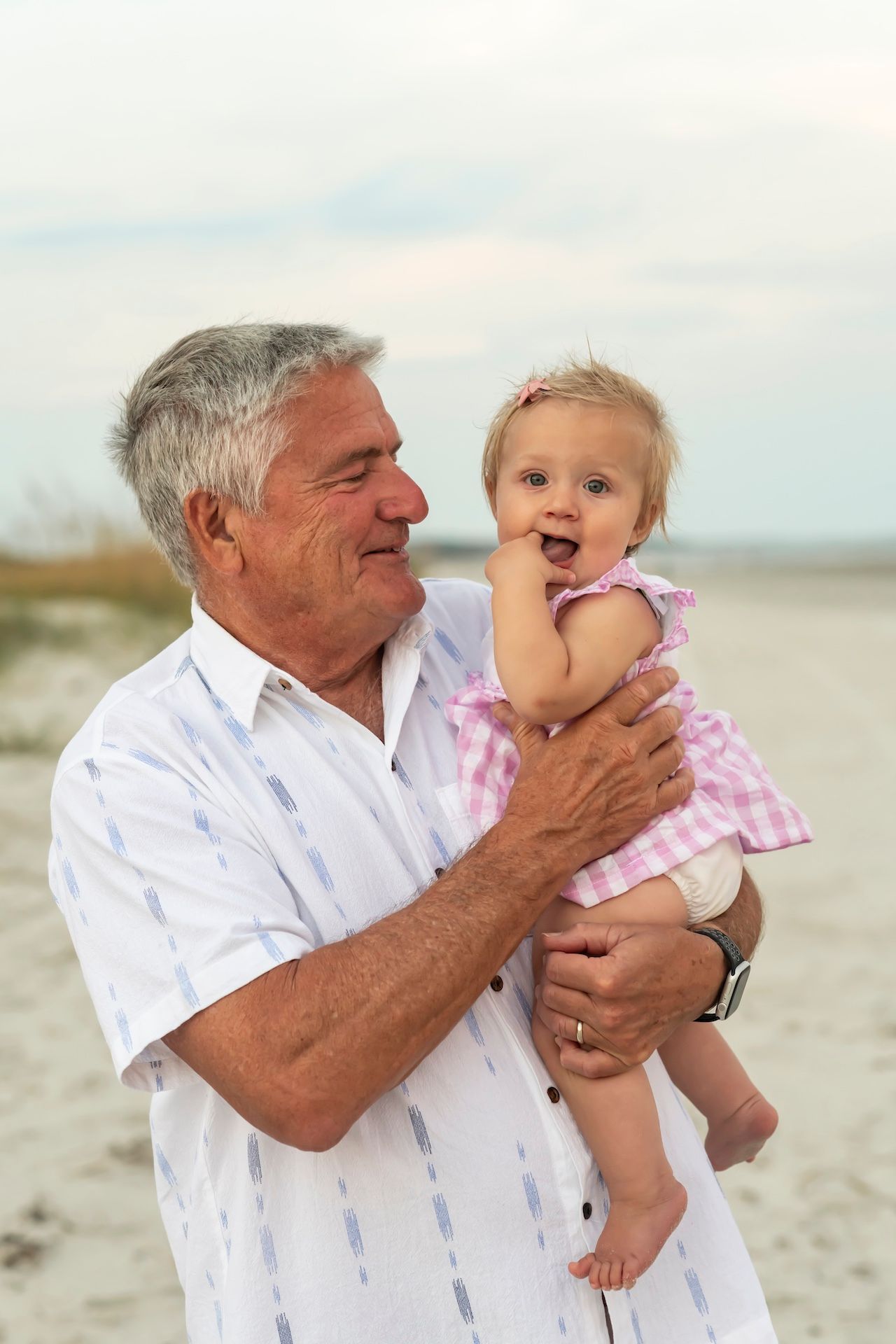 Joyful family moment at beach as senior man in white shirt holds excited baby in pink dress.