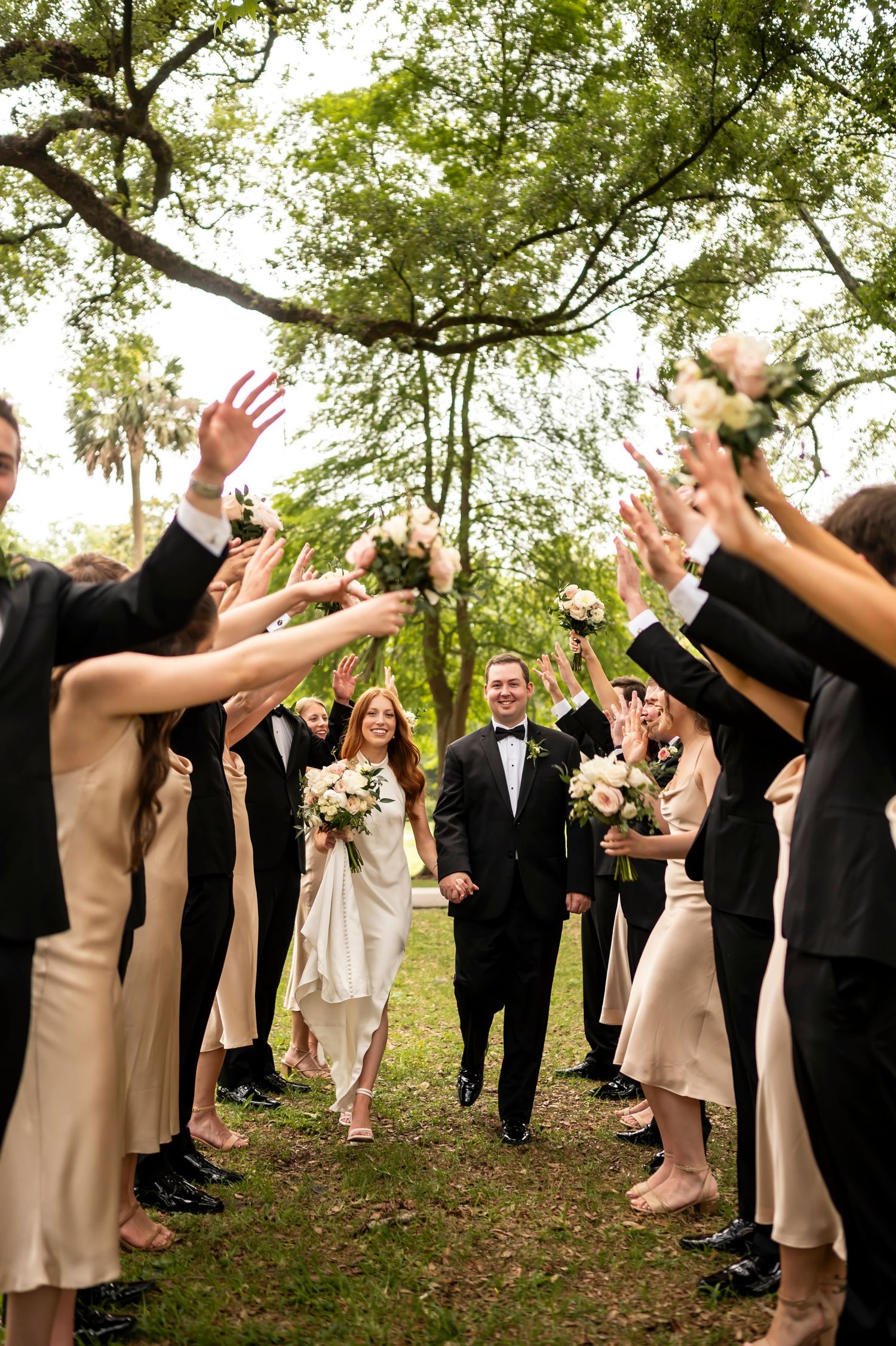 Wedding party members in formal attire throw their hands up joyfully during outdoor celebration in wooded setting.
