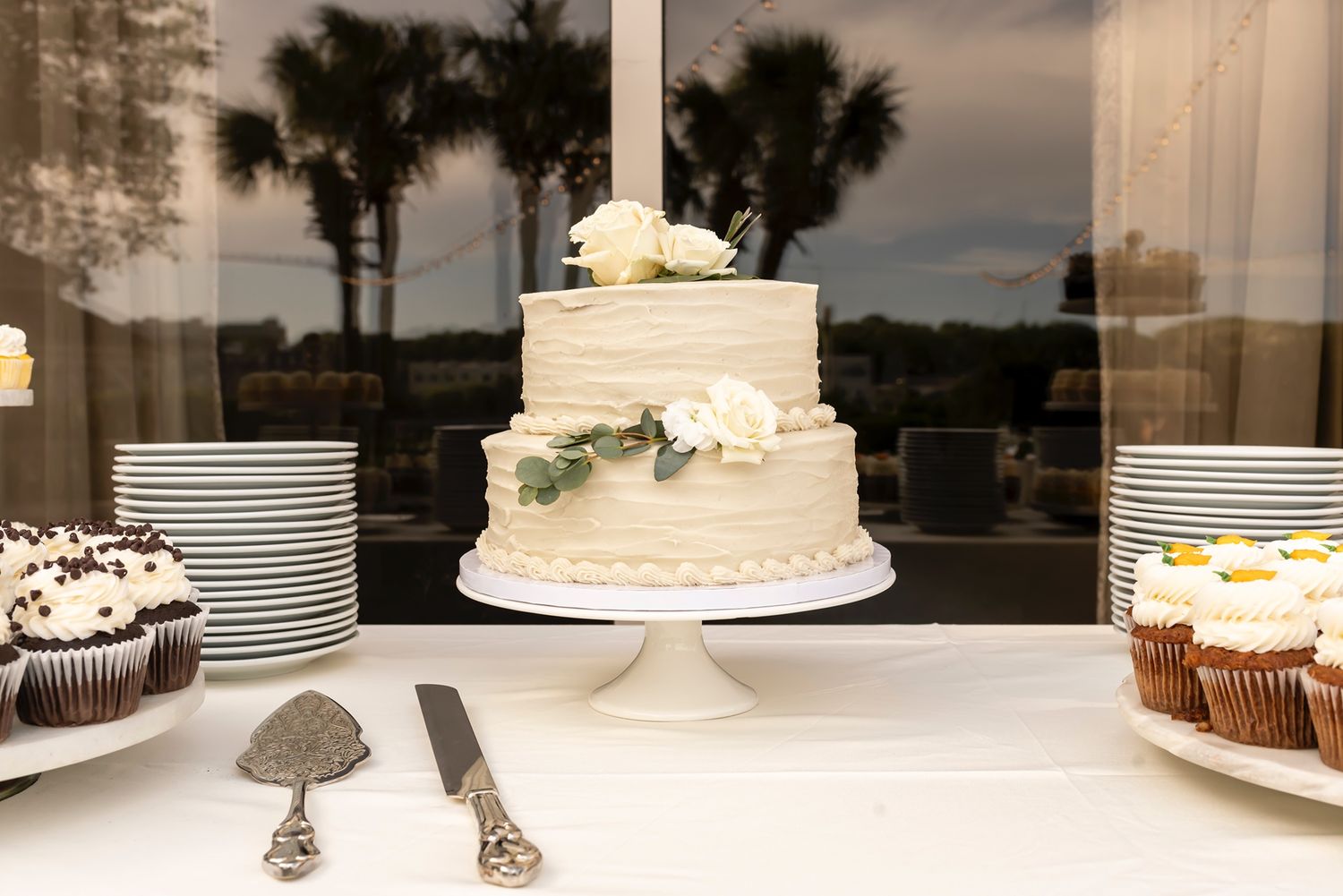 A two-tiered white wedding cake on a pedestal surrounded by cupcakes and place settings with palm trees visible outside.