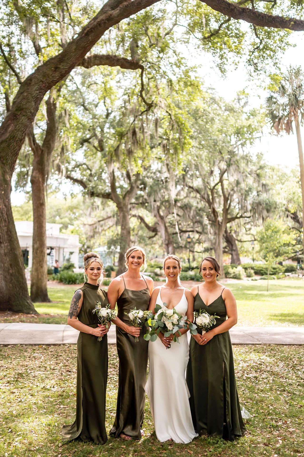 A group of bridesmaids in elegant olive green gowns pose with a bride under Spanish moss draped oak trees.