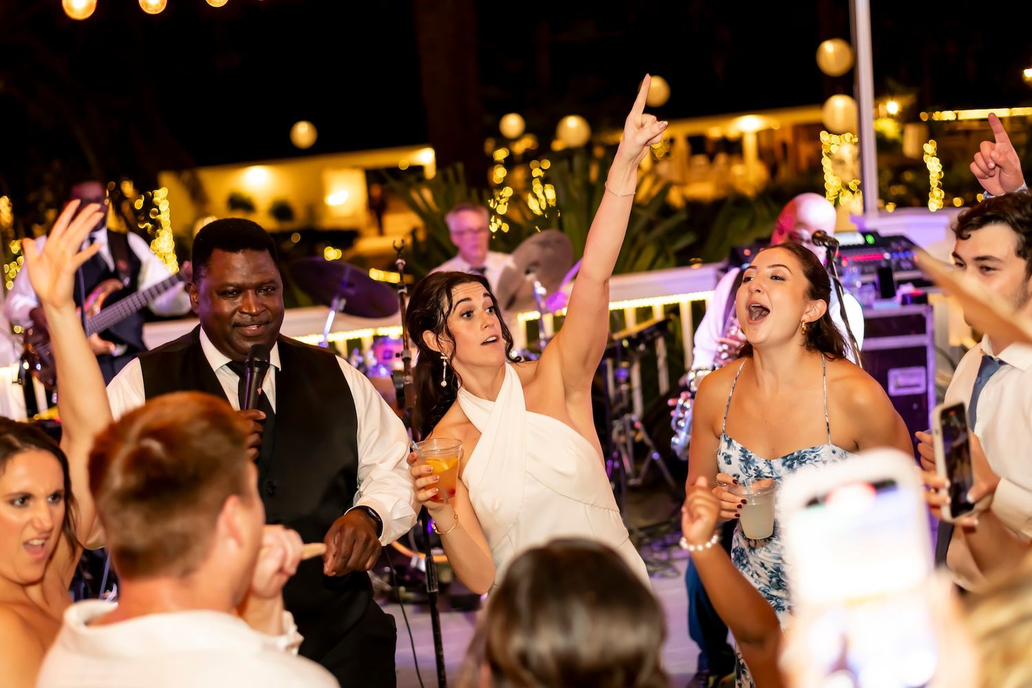 Wedding party guests dance and celebrate enthusiastically during an evening reception with glowing lights in the background.