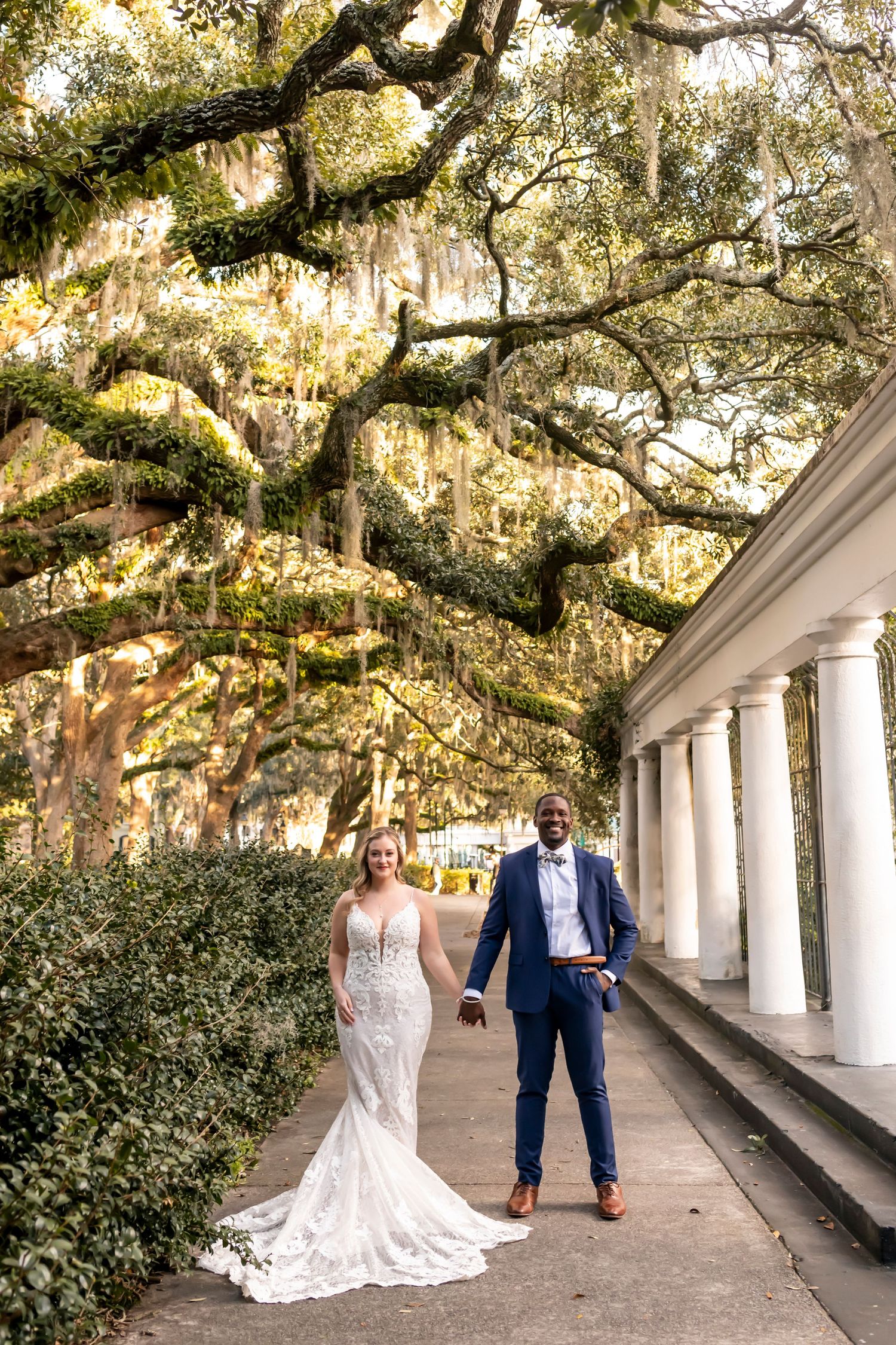 A newly married couple poses beneath Spanish moss-draped oak trees along a columned walkway in the Southern style.