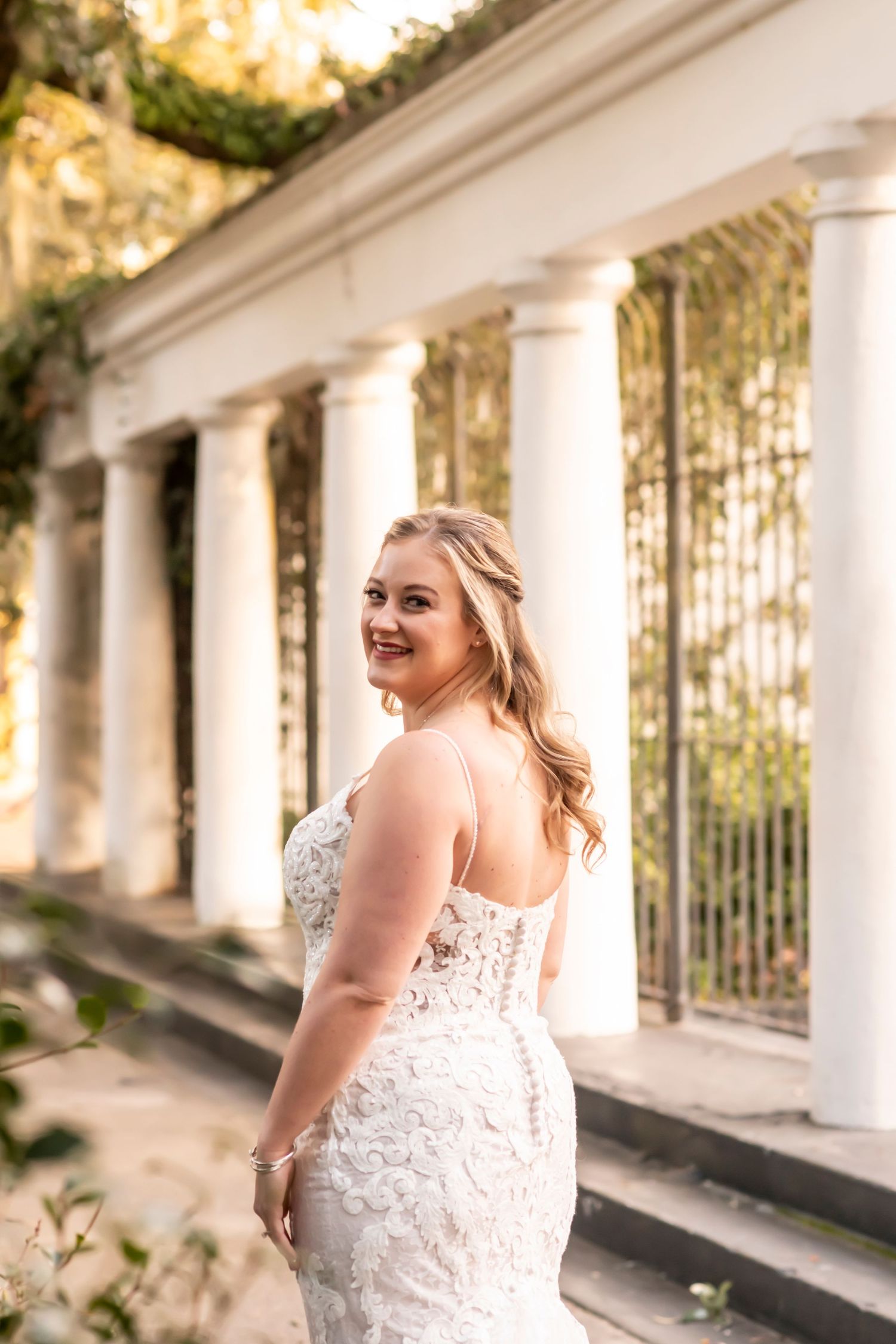 A bride in a fitted lace wedding dress poses by classical white columns in a garden setting during golden hour.
