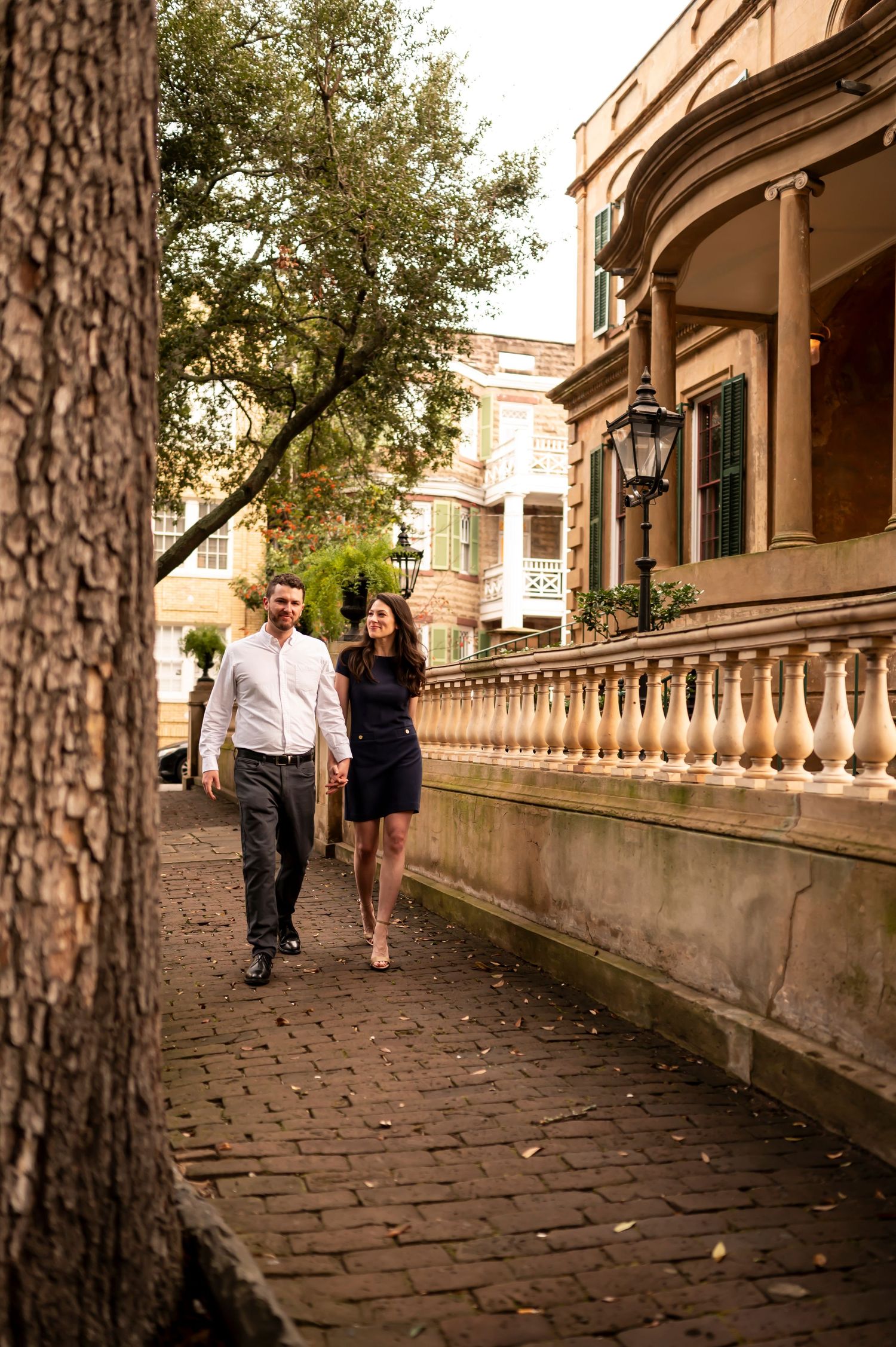 A couple walks along a stone pathway bordered by ornate balusters outside a historic building at sunset.