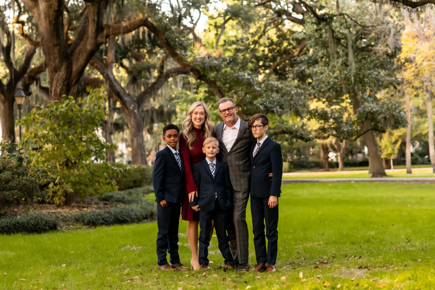 A group of people in formal attire stand together on lush green grass near trees in an outdoor park setting.