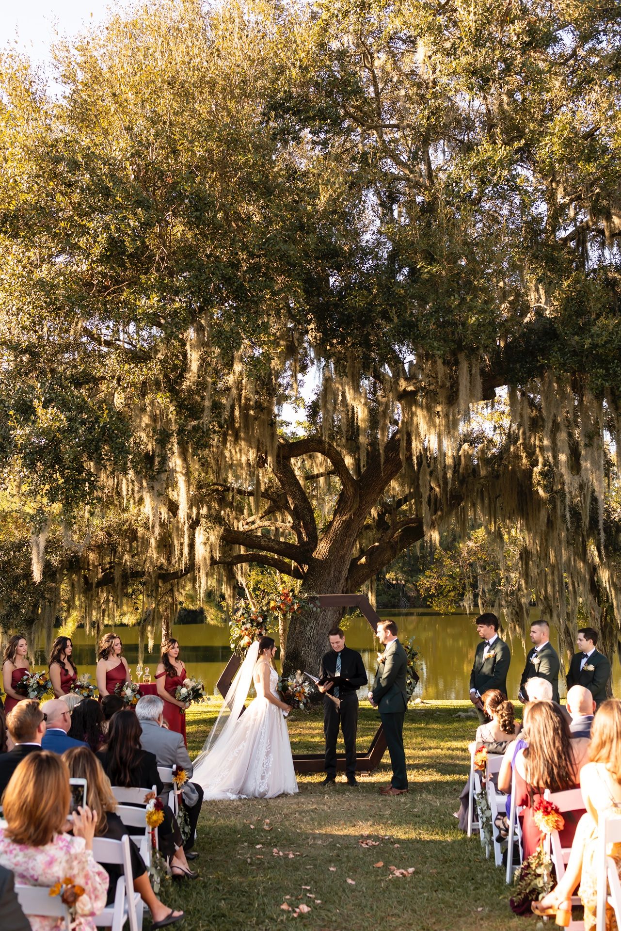 An outdoor wedding ceremony takes place under moss-draped oak trees as guests watch from white chairs.