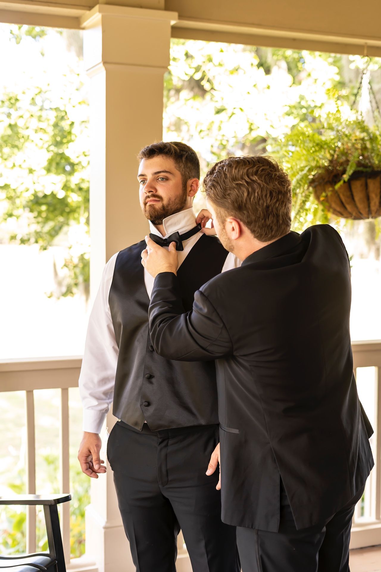 Someone helps adjust another person's bowtie on a porch with hanging plants in the background.