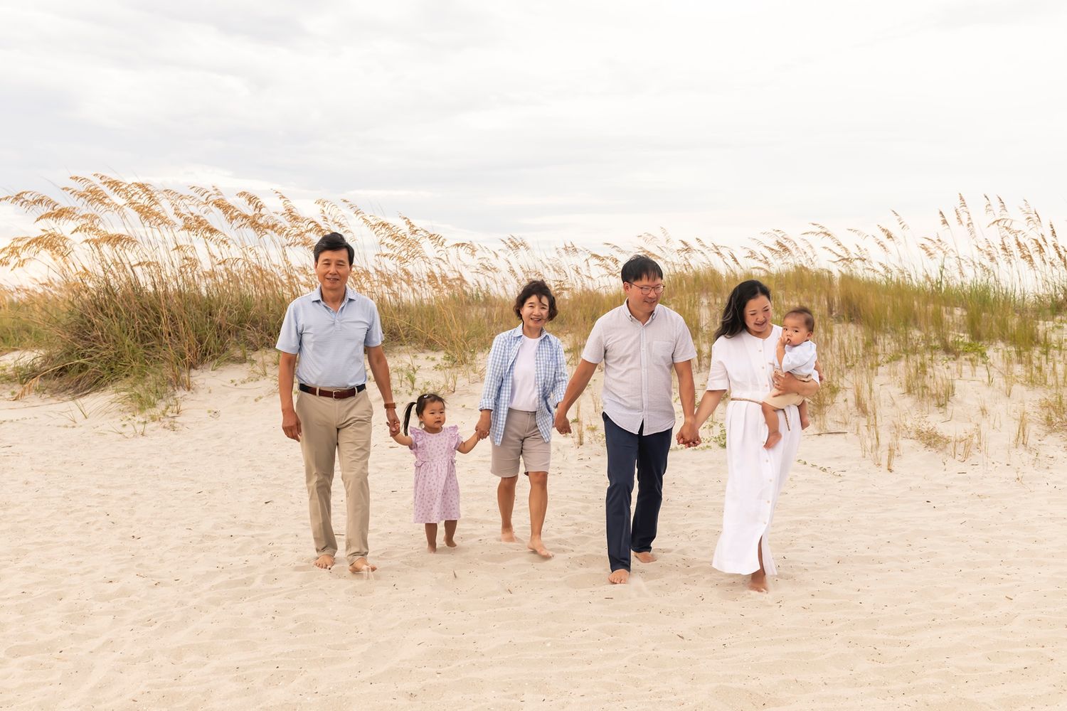 A group of people walk hand in hand along a sandy beach path lined with tall coastal grasses at sunset.