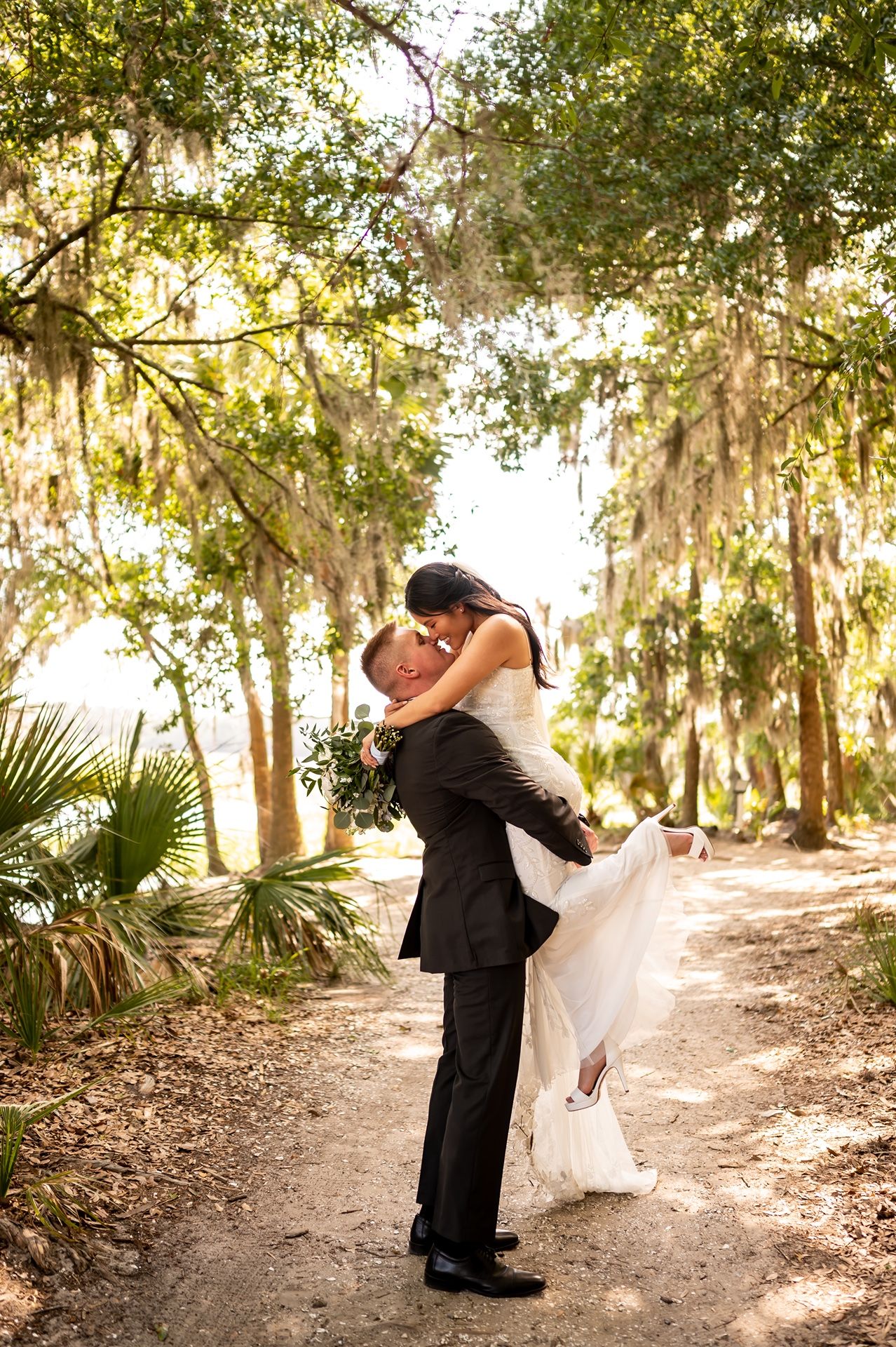 A couple shares a romantic moment on a sunlit forest path lined with Spanish moss and palm trees.