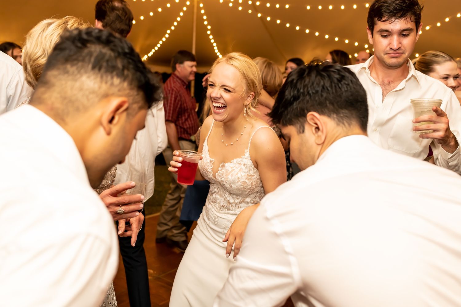 A bride in a white dress dances and laughs with wedding guests under string lights at an indoor reception.