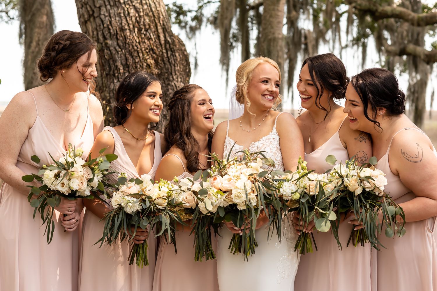 A group wearing matching blush pink bridesmaid dresses laughing together while holding floral bouquets.