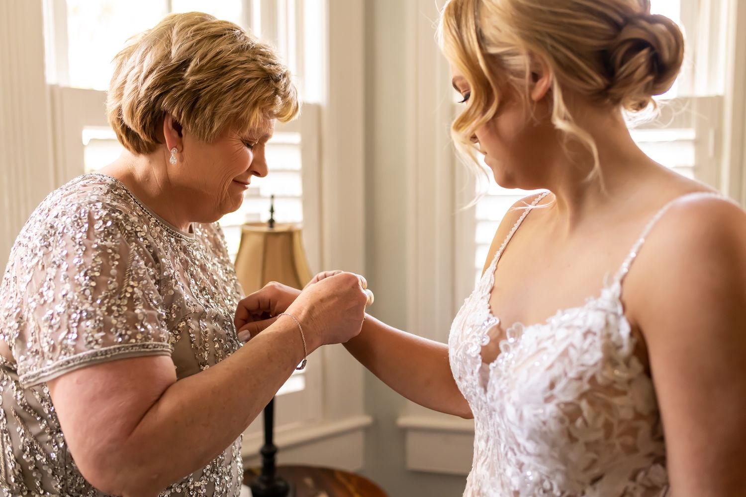 Two people share an intimate moment in a sunlit room as someone helps adjust a delicate white lace dress.