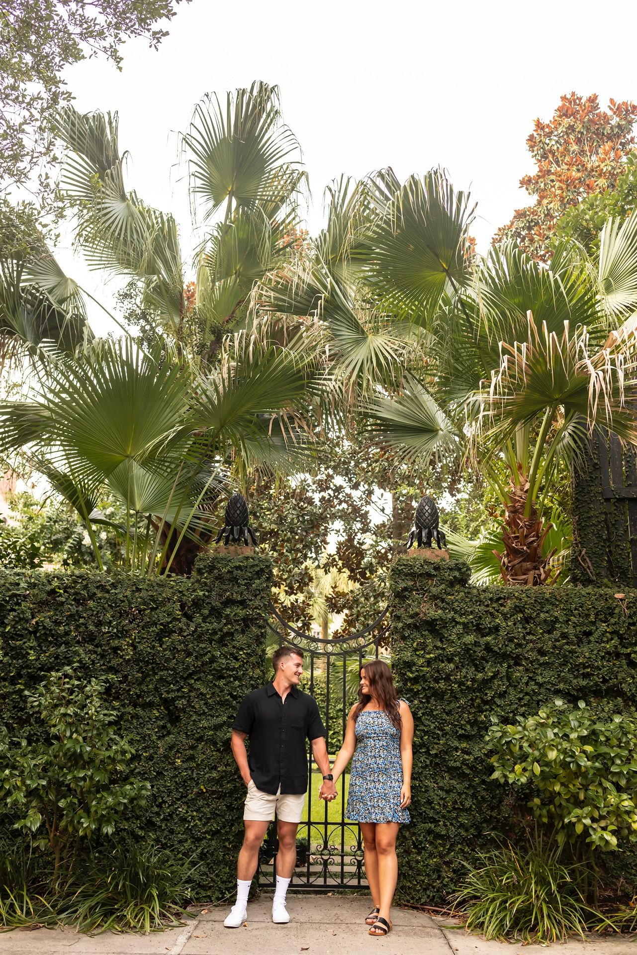 Palm trees and lush greenery line a garden wall while two people hold a sign outdoors during daytime.