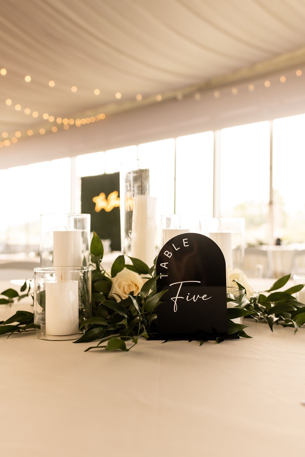 Black heart-shaped table number displaying 'Table Five' with white candles and greenery at an elegant wedding reception.